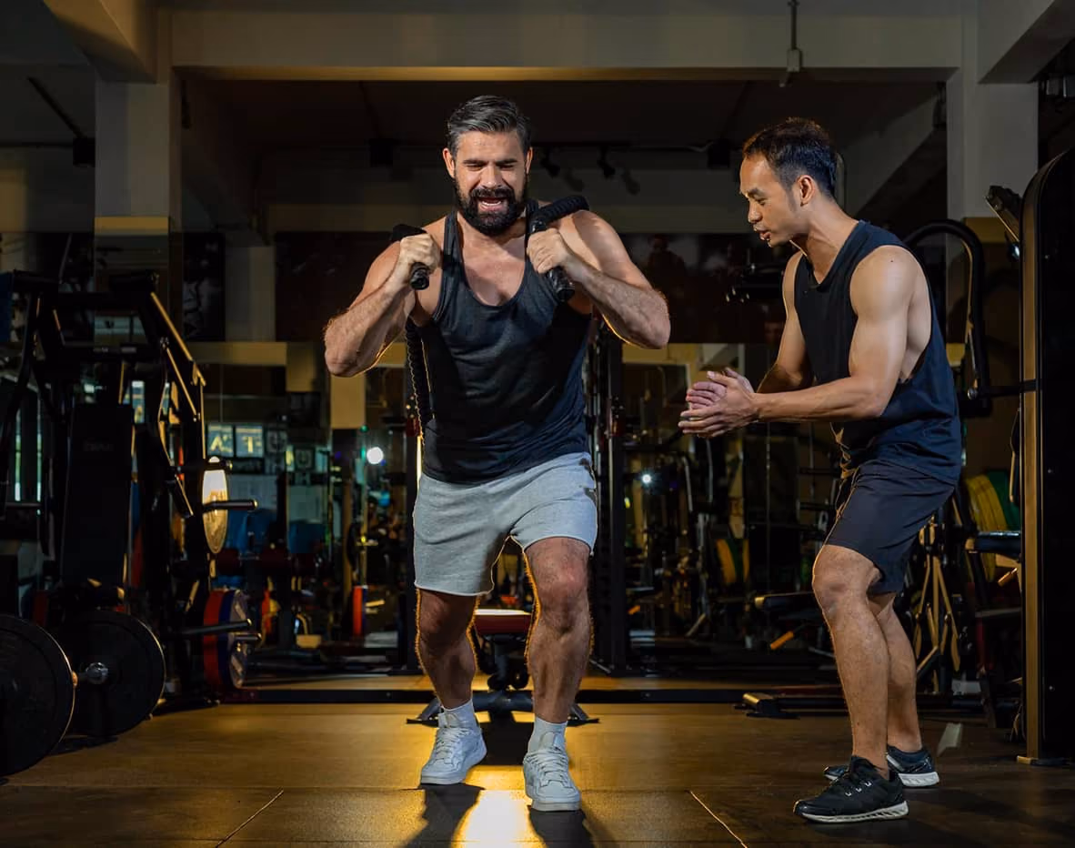 Man in gray shorts and black tank top working out with resistance bands while trainer encourages him.