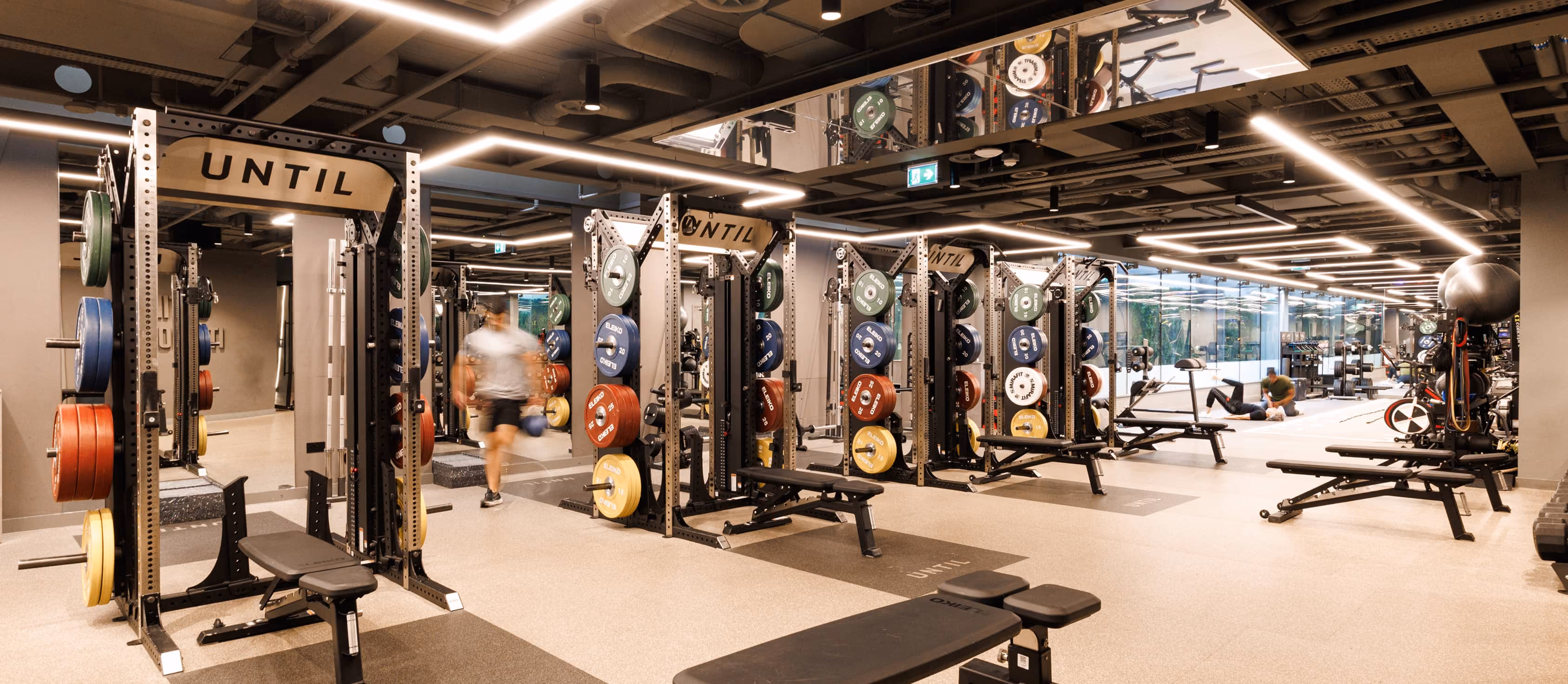 Modern gym with multiple weight racks, benches, bright lighting, and two people exercising in background.