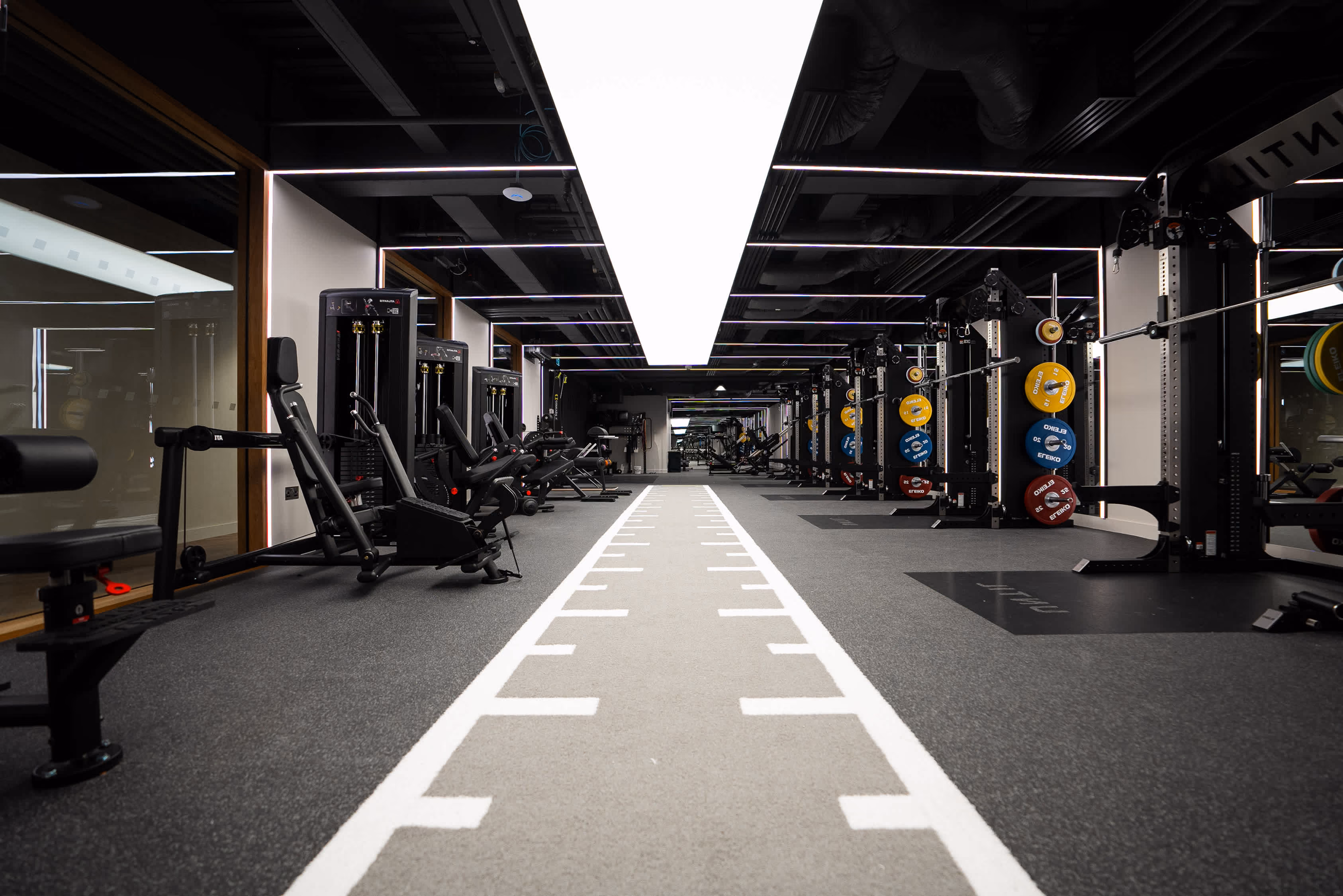 Modern gym interior with weight machines, racks, and a white-lined turf strip in the center.