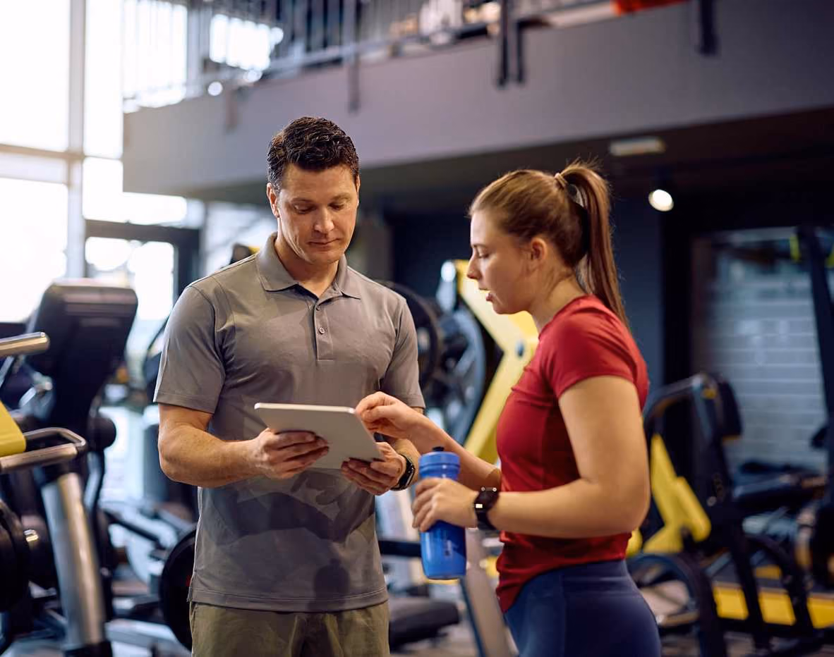 Gym trainer showing a tablet to a female client holding a water bottle inside a fitness center.