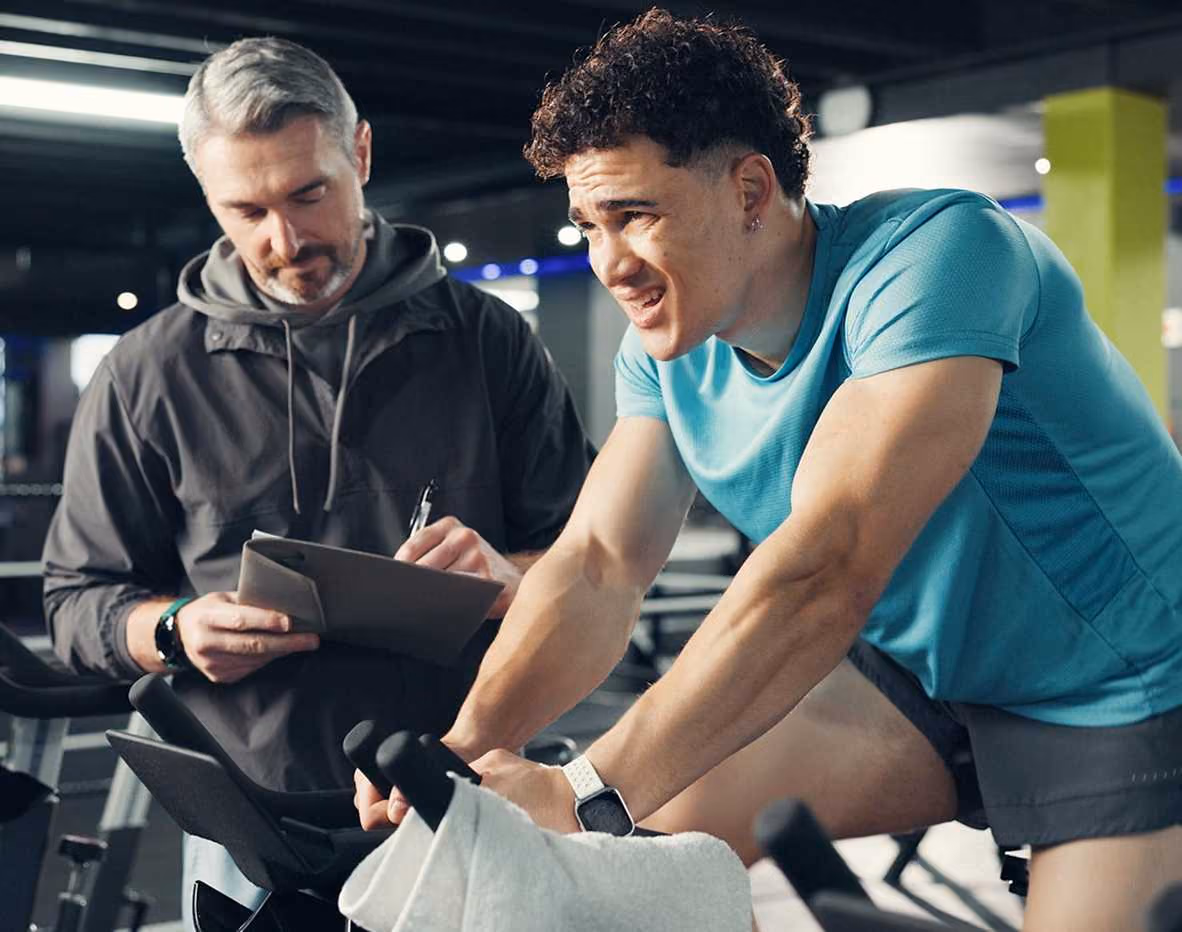 Trainer recording data while athlete exhausts on stationary bike in gym with towel draped over handlebars.