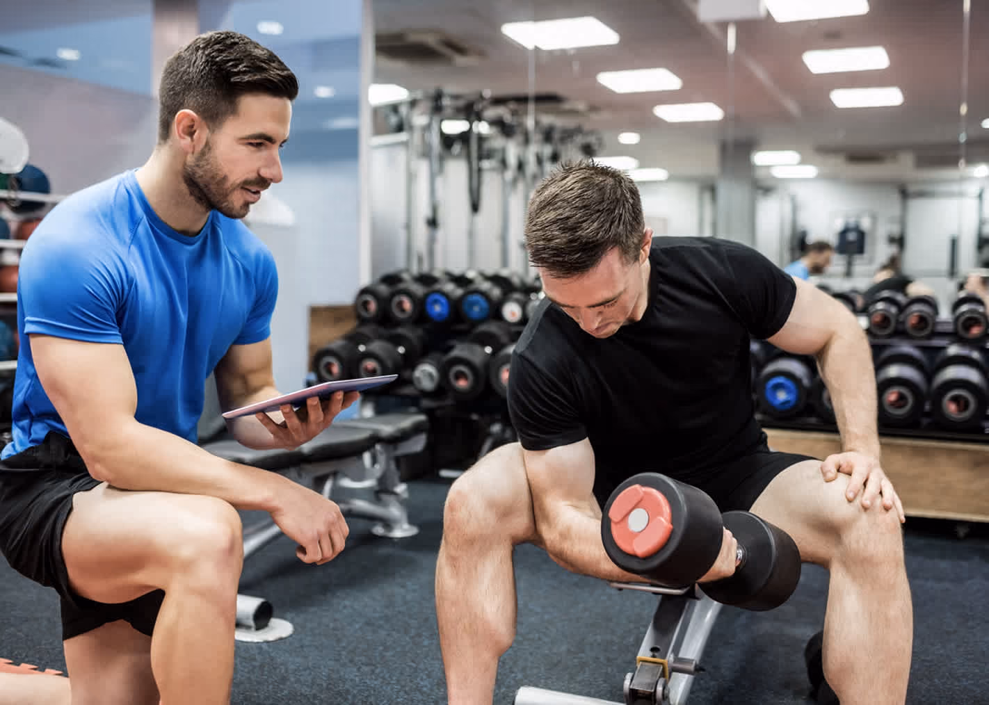 Man lifting a dumbbell on a bench as a trainer watches holding a tablet in a gym.