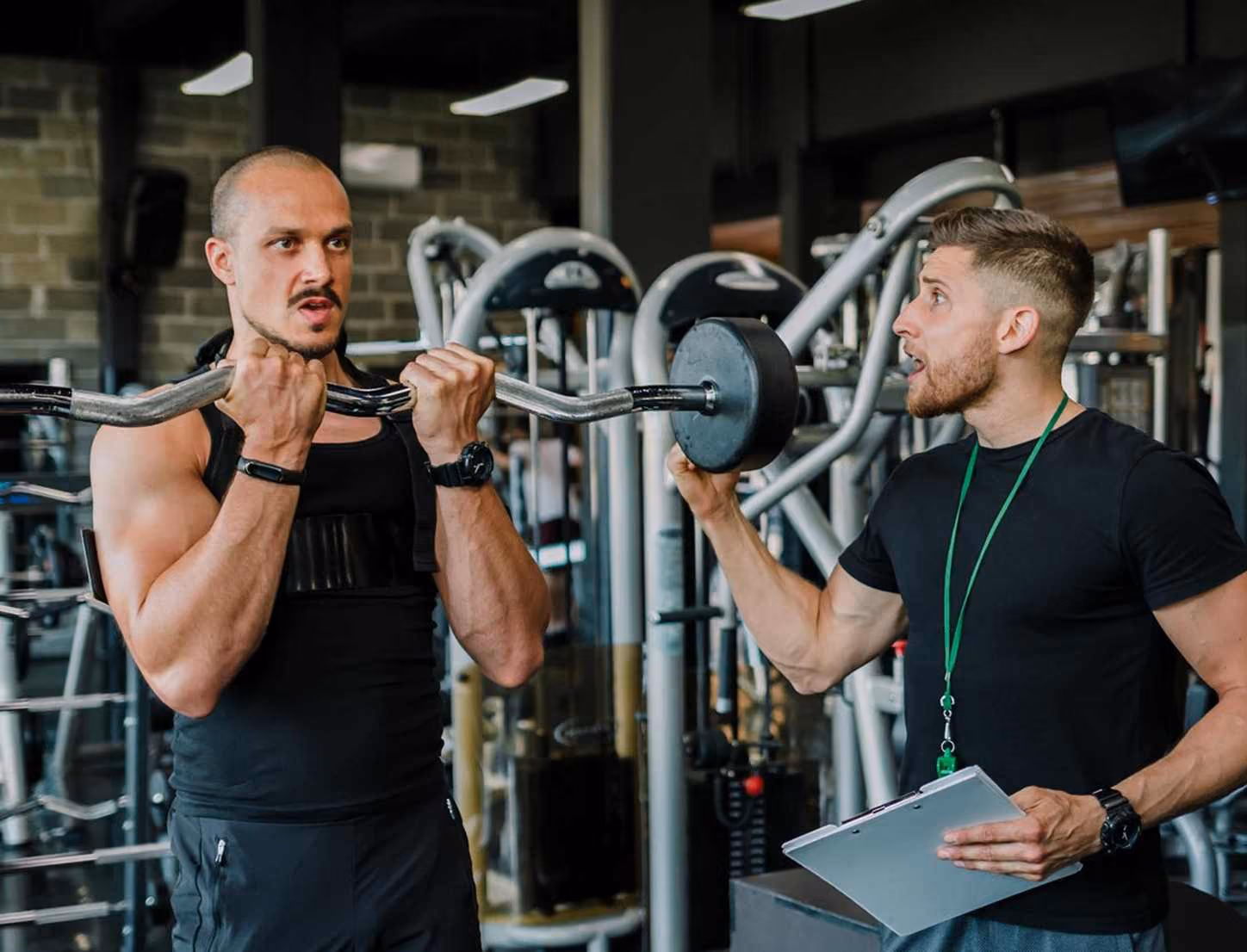Man lifting barbell curls while trainer coaches and holds clipboard in gym setting.