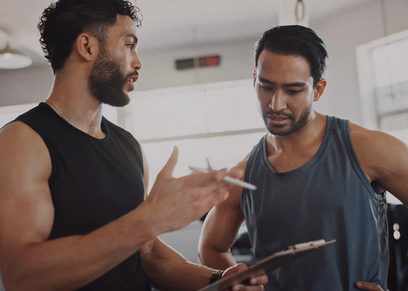 Two muscular men in gym tank tops discussing notes on a clipboard during a workout session.