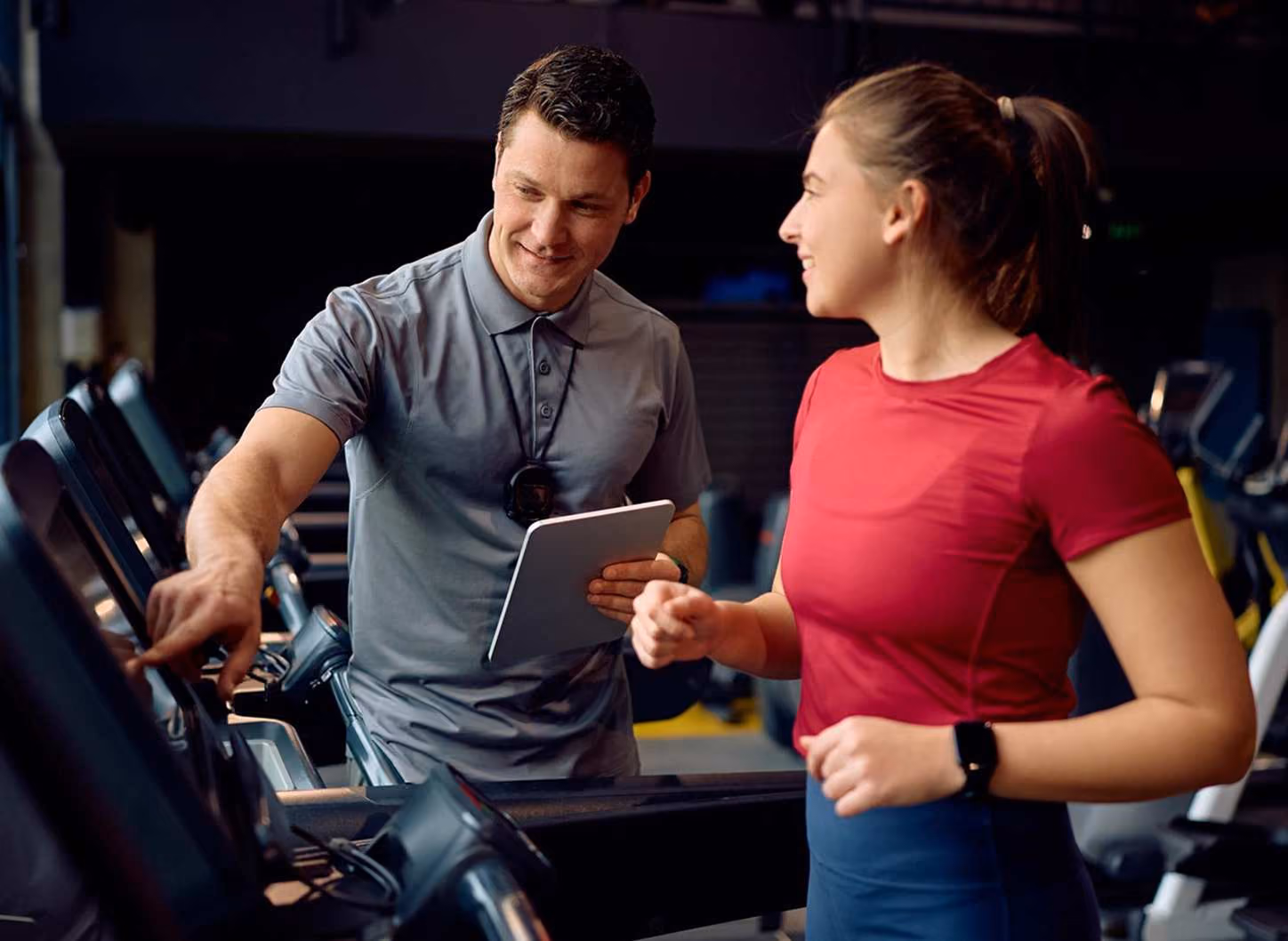 Trainer with stopwatch and tablet guiding woman in red shirt on treadmill at gym.