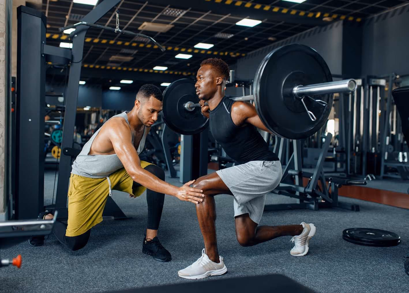 Trainer assisting man doing weighted lunges in gym with barbell on shoulders.