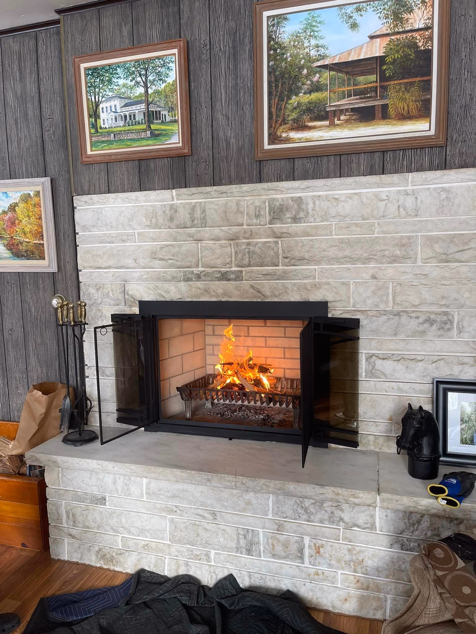 Stone fireplace with glass doors open and fire burning, surrounded by paintings and decorative items.