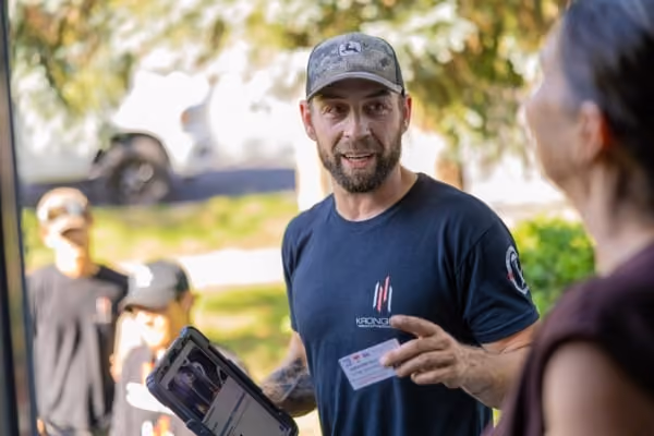 Chimney worker wearing a cap and black shirt holding a tablet and ID card outdoors, talking to a woman.