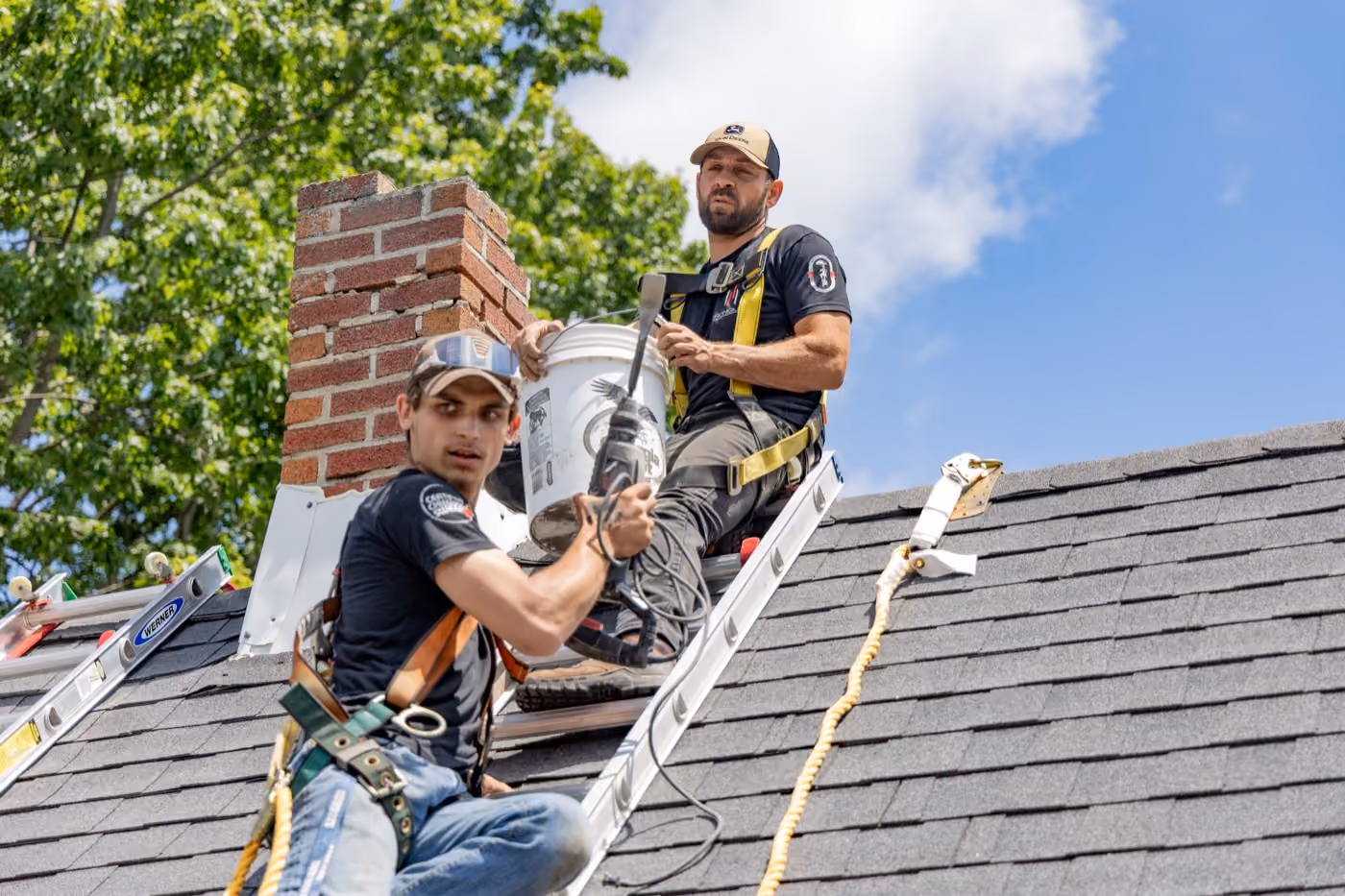 Two chimney workers on a roof repairing a brick chimney with safety harnesses and tools, trees and sky in background.