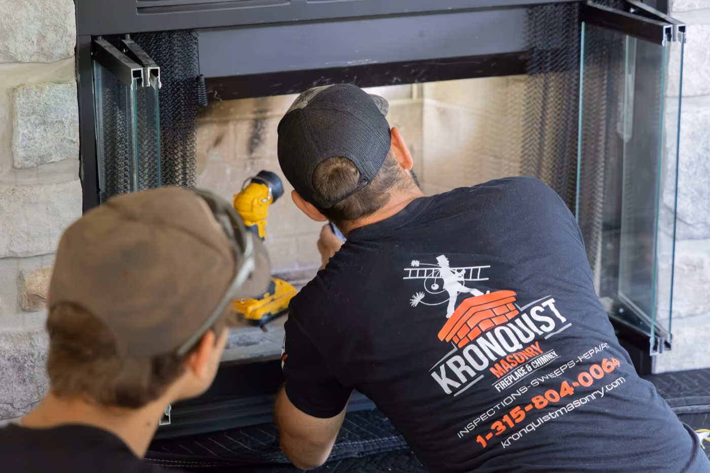 Two men working on a fireplace, one wearing a black shirt with Kronquist Masonry logo and contact info.