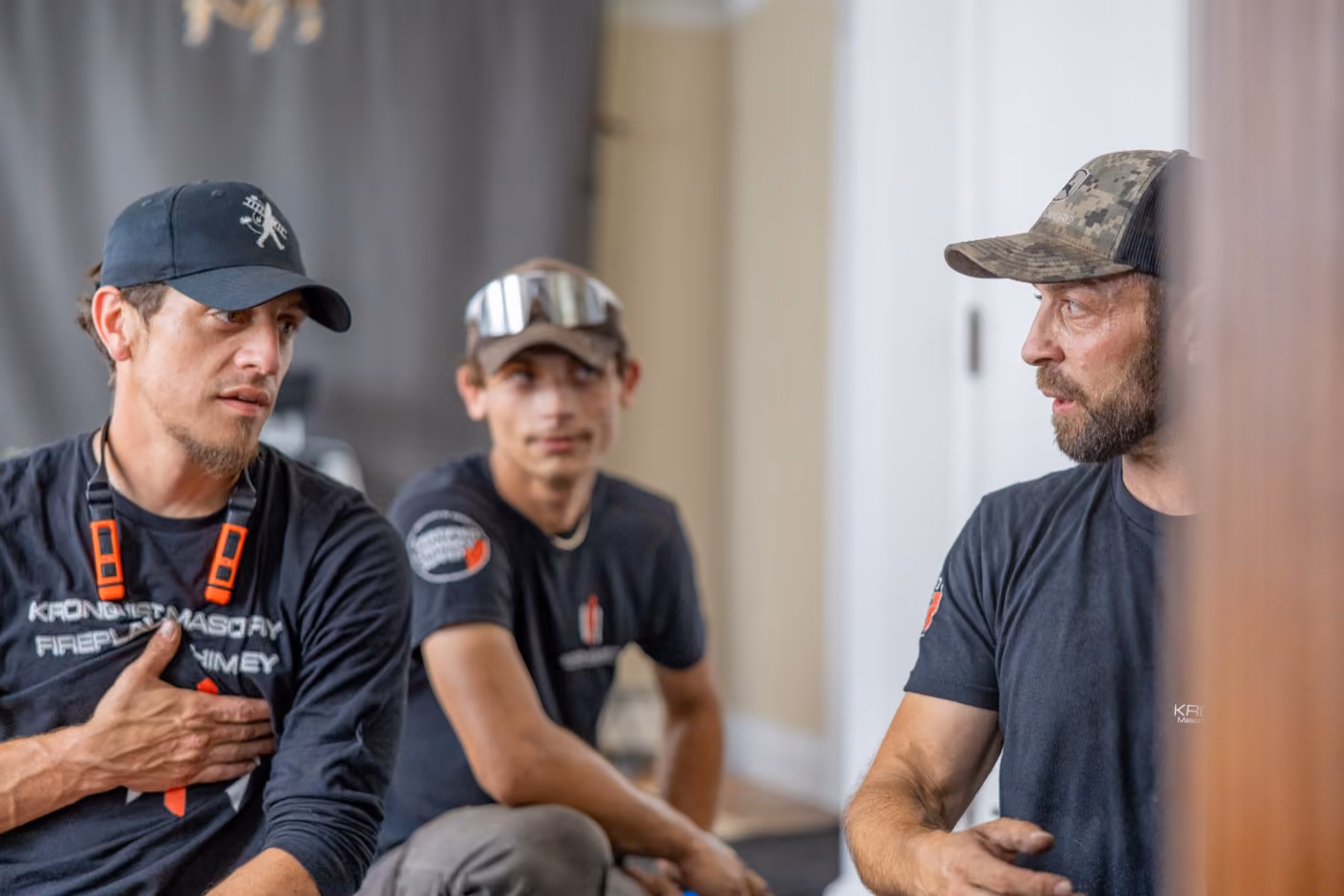 Three chimney workers in casual work clothes and caps engaged in conversation indoors.