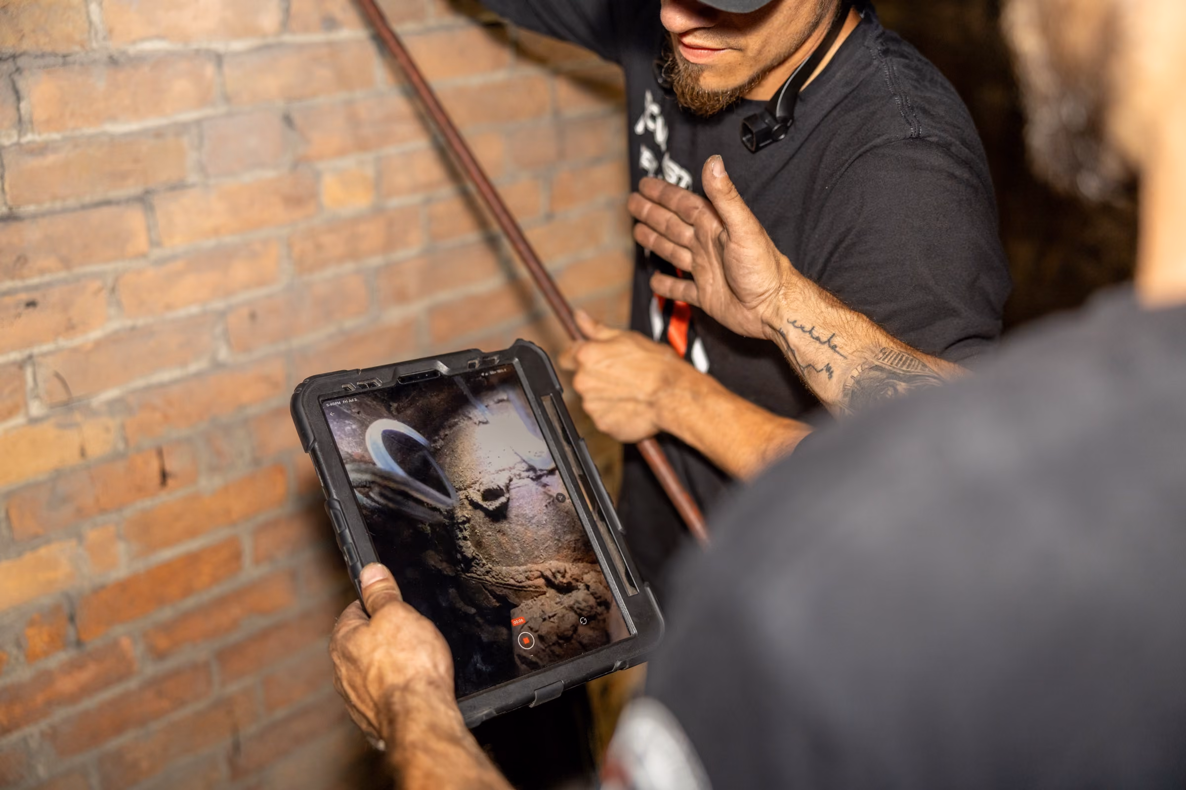 Two men inspecting chimney footage on a tablet inside a brick-walled area.