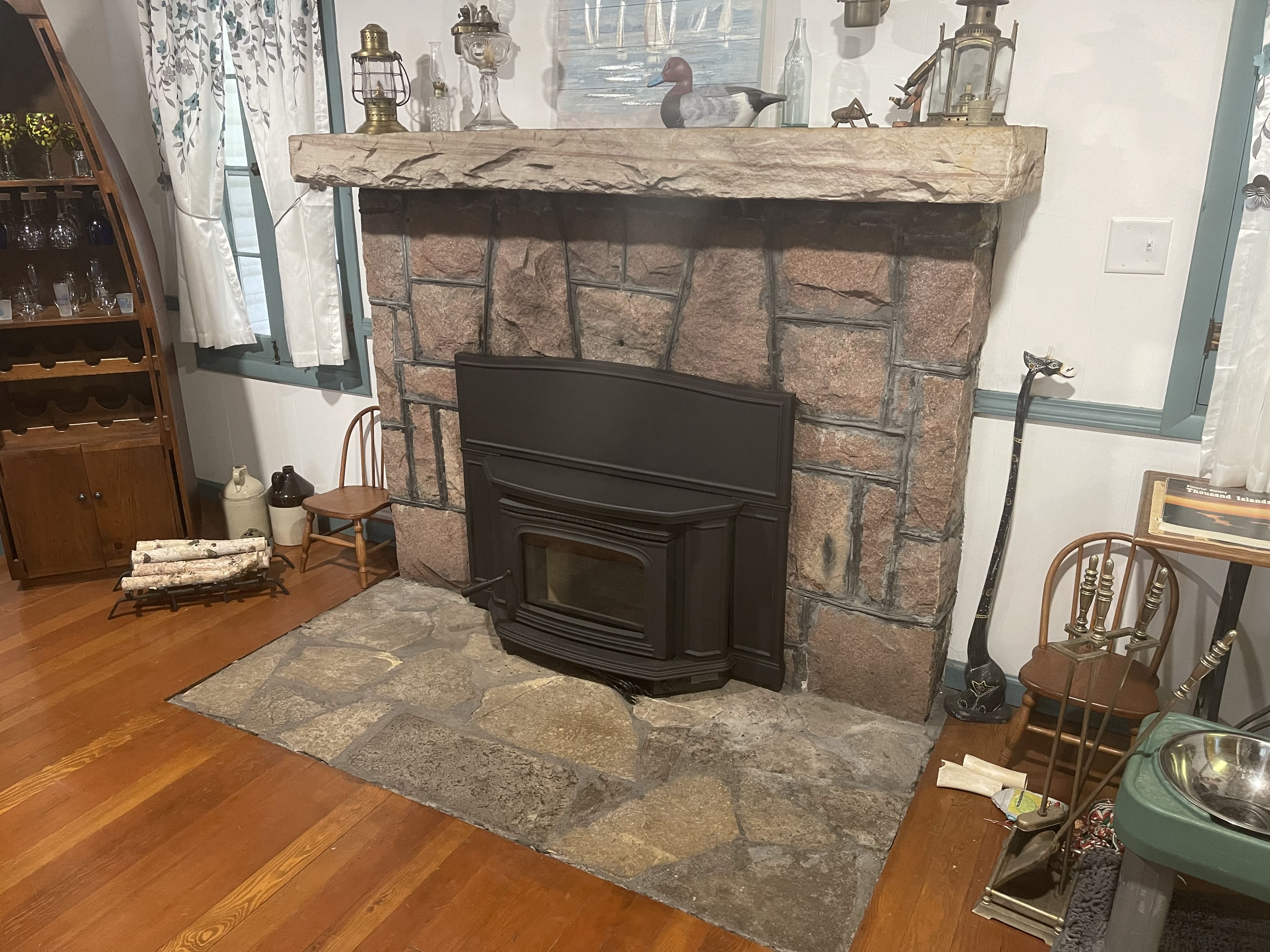 Stone fireplace with black insert surrounded by wood floor and rustic decor including chairs and lanterns.