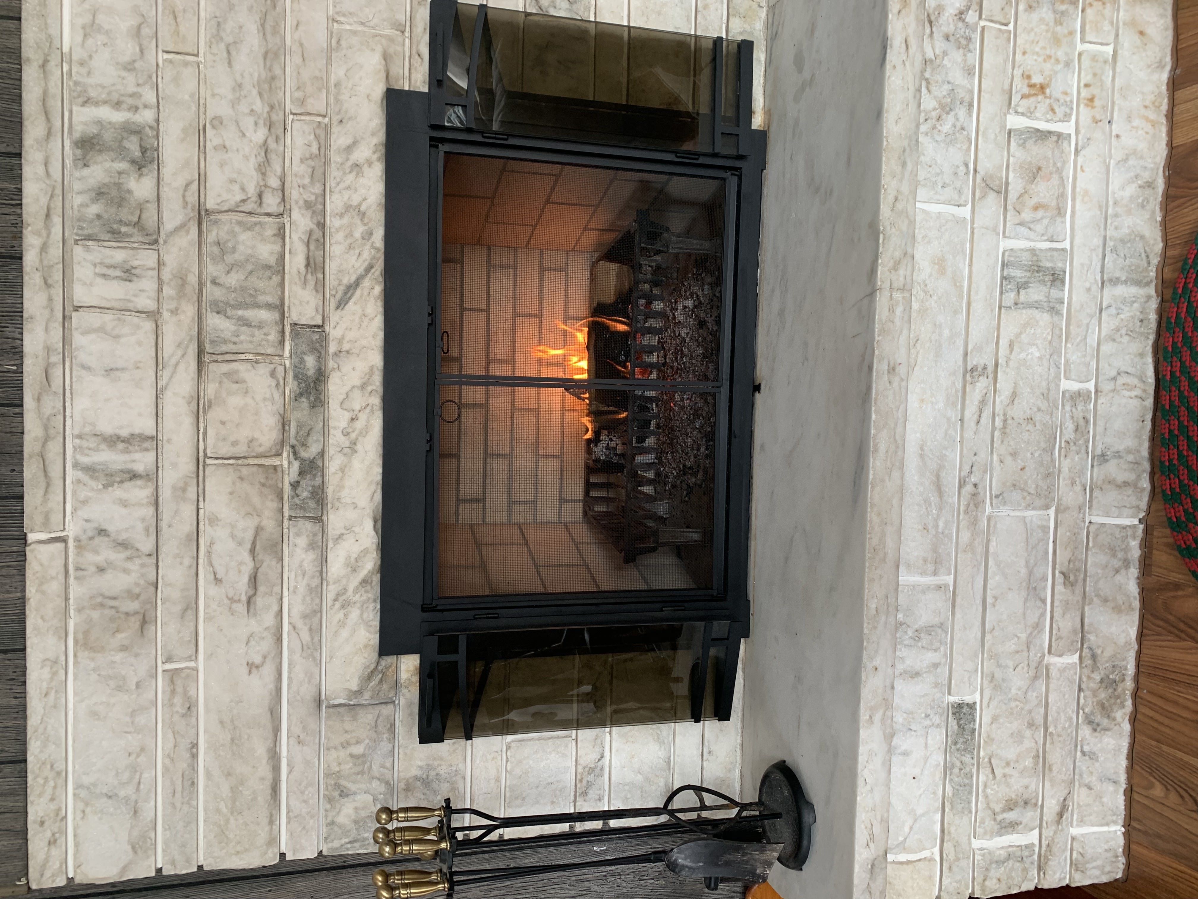Lit fireplace with glass doors set in light stone surround and black metal frame with fireplace tools nearby.