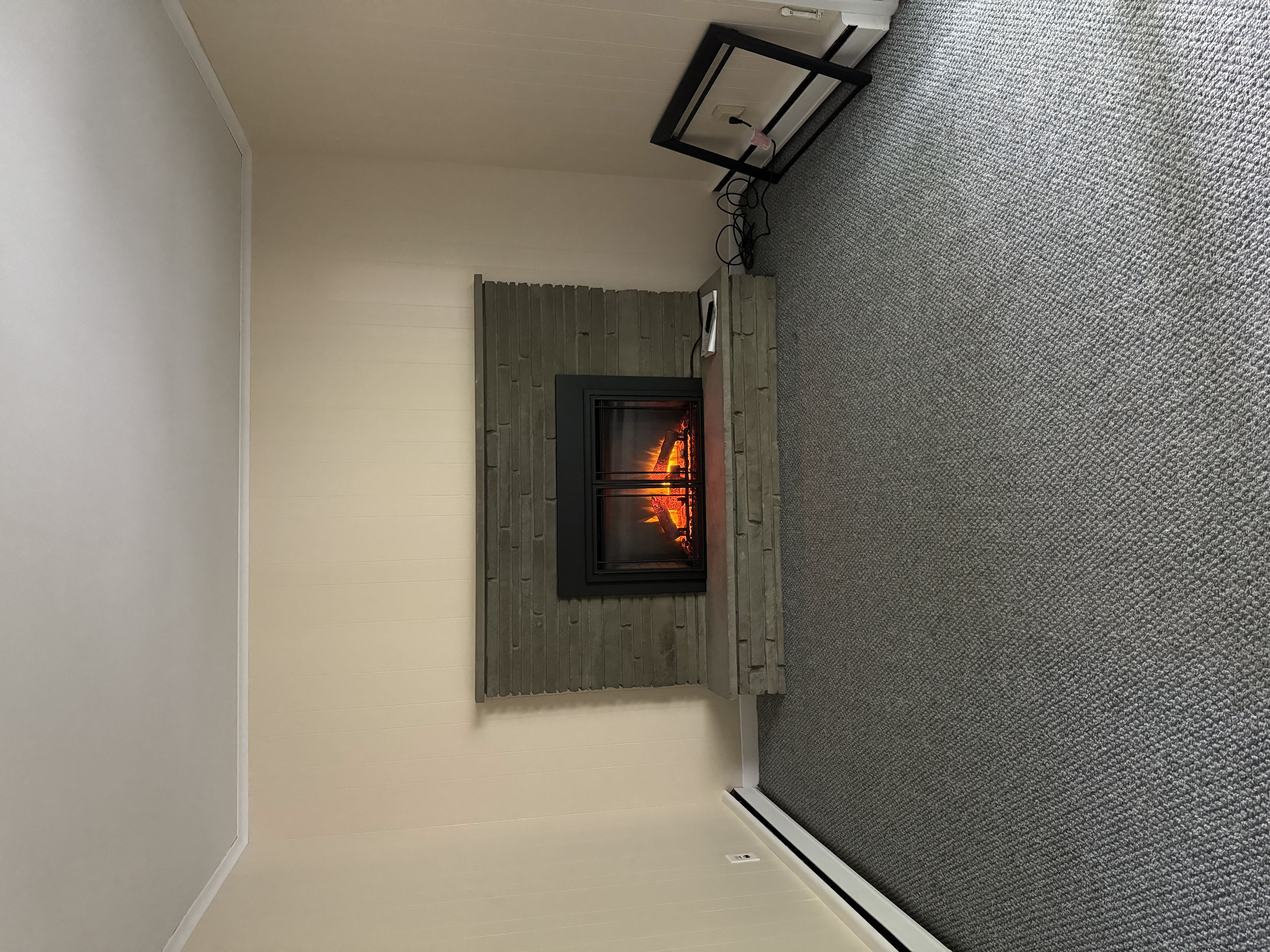 Empty room with gray carpet, beige walls, and a stone fireplace with a fire burning inside.