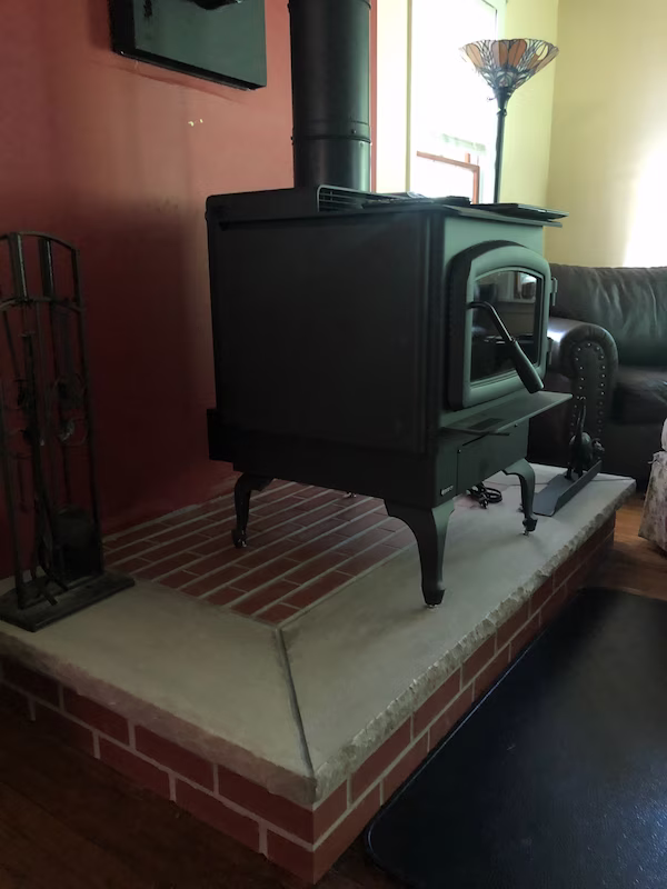 Black wood stove with glass door on a brick and concrete hearth in a cozy living room.