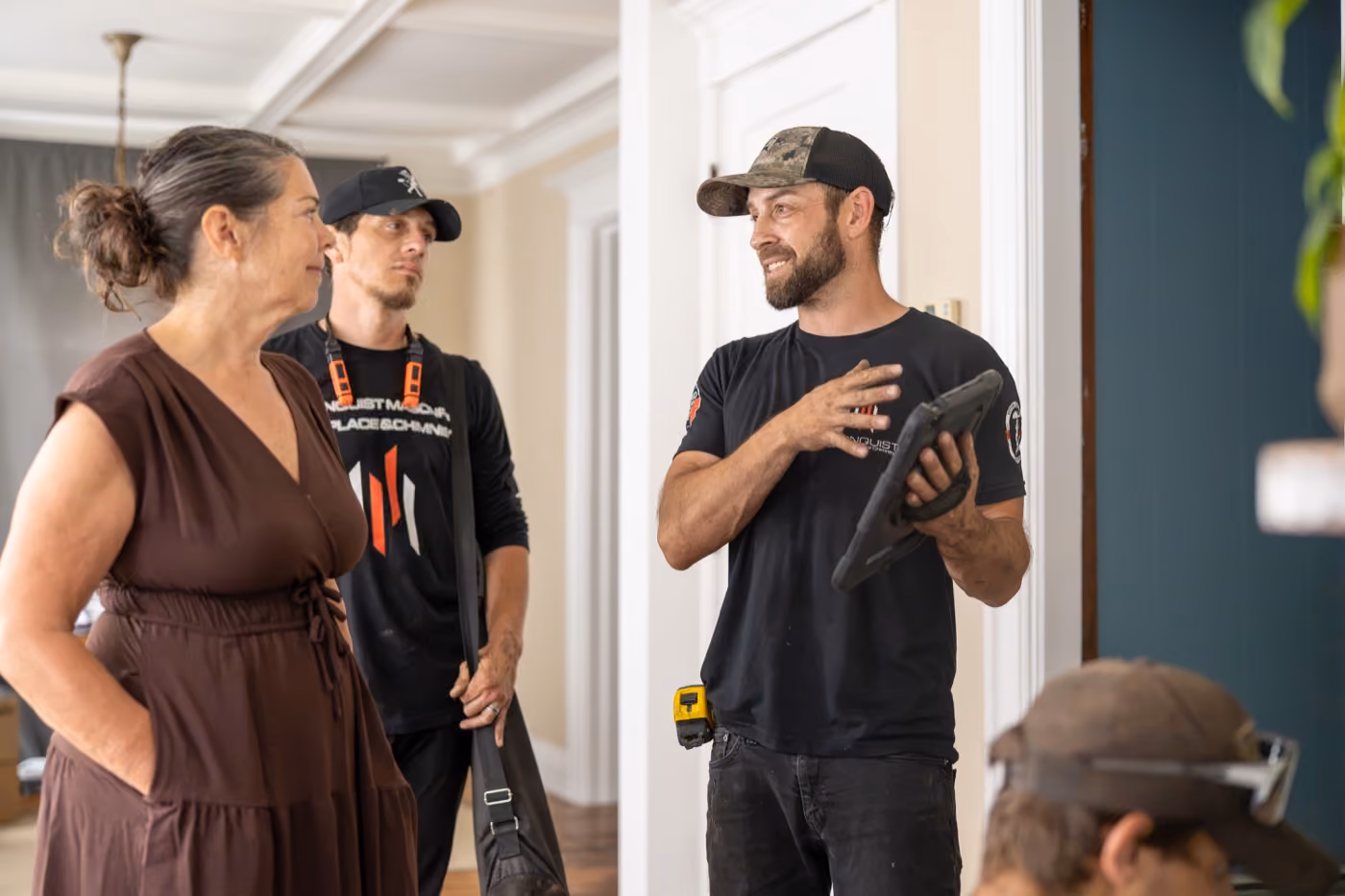 Two chimney technicians in work shirts and caps talk with a woman in a brown dress inside a home.