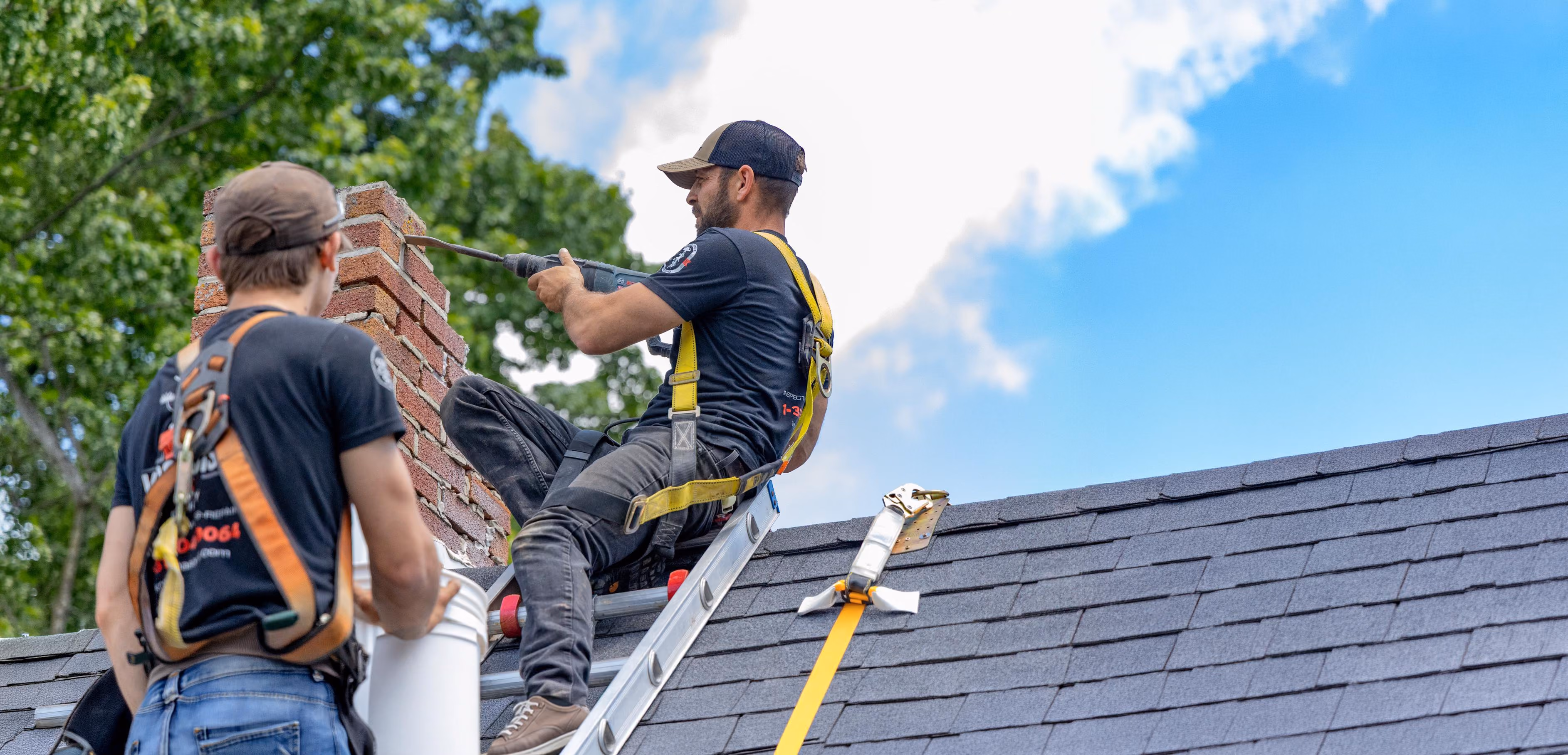 Two workers in harnesses repairing a brick chimney on a roof under a blue sky.