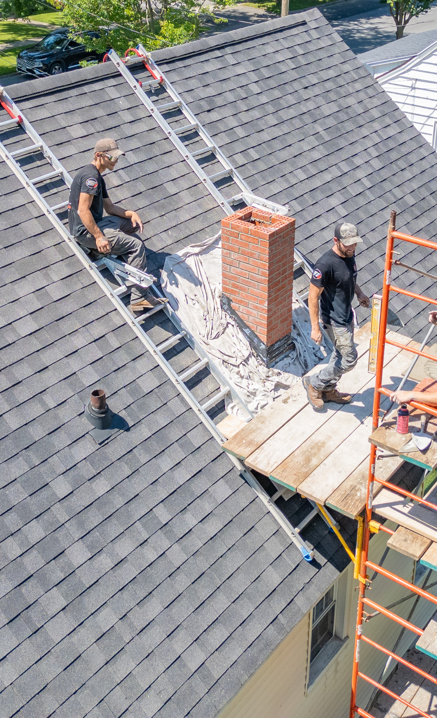Two workers in harnesses repairing a brick chimney on a roof under a blue sky.