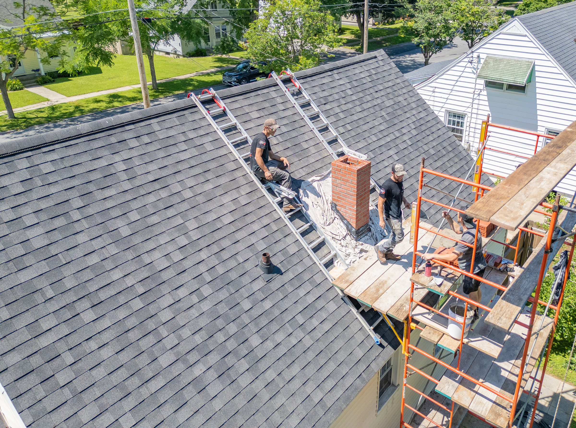 Two workers in harnesses repairing a brick chimney on a roof under a blue sky.