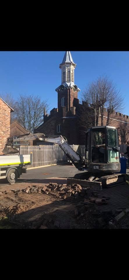 Small excavator clearing bricks on a paved area with a brick building and steeple in the background.