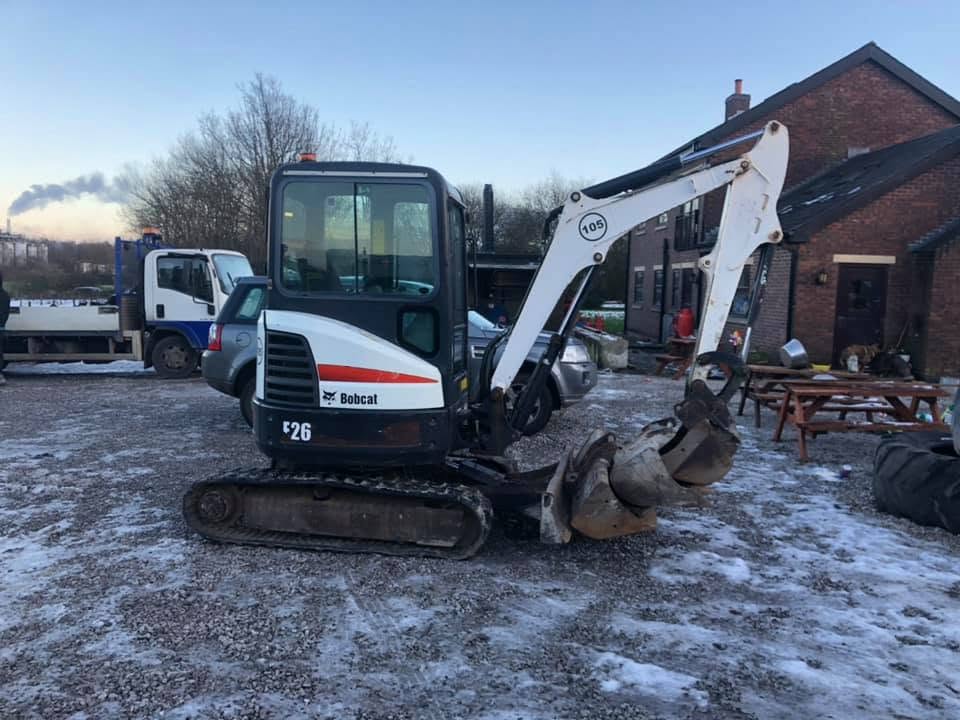 White Bobcat compact excavator with dual buckets on snowy ground near brick building and vehicles.