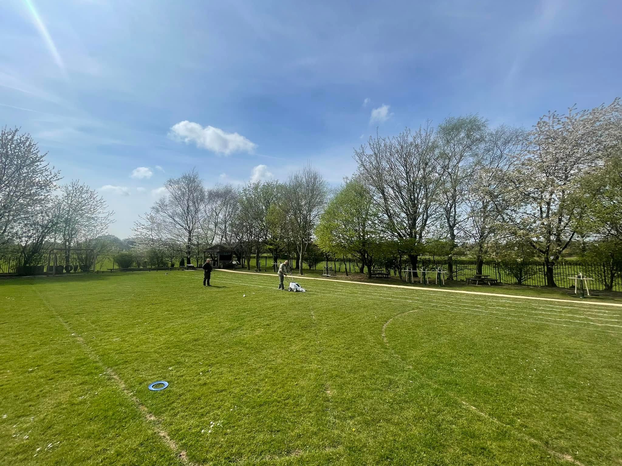 Two people marking lines and mowing grass on a green grass field with trees and blue sky in the background.
