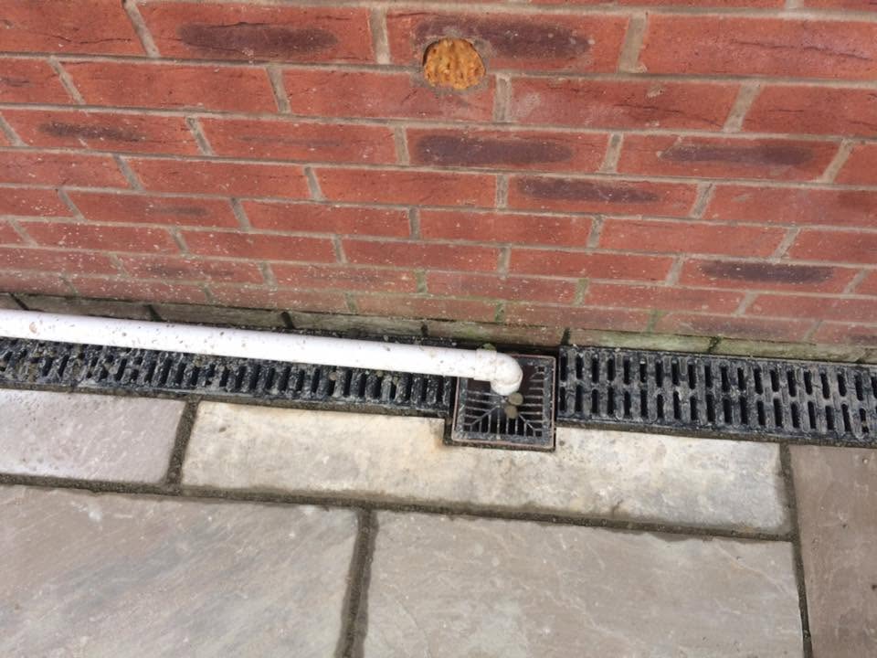 French drain running alongside a house between a red brick wall and some flagging.
