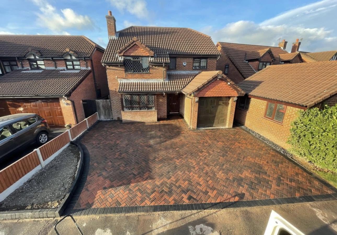 Brick driveway with a black border in front of a two-story house with a garage and brown tiled roof.