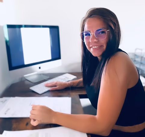 Frau mit Brille sitzt lächelnd an einem Schreibtisch vor einem Computer mit Dokumenten.