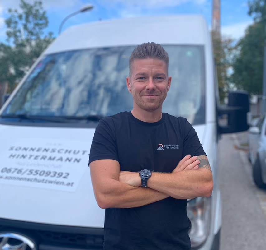 Ein Mann in schwarzem T-Shirt mit verschränkten Armen vor einem weißen Lieferwagen mit Firmenbeschriftung.