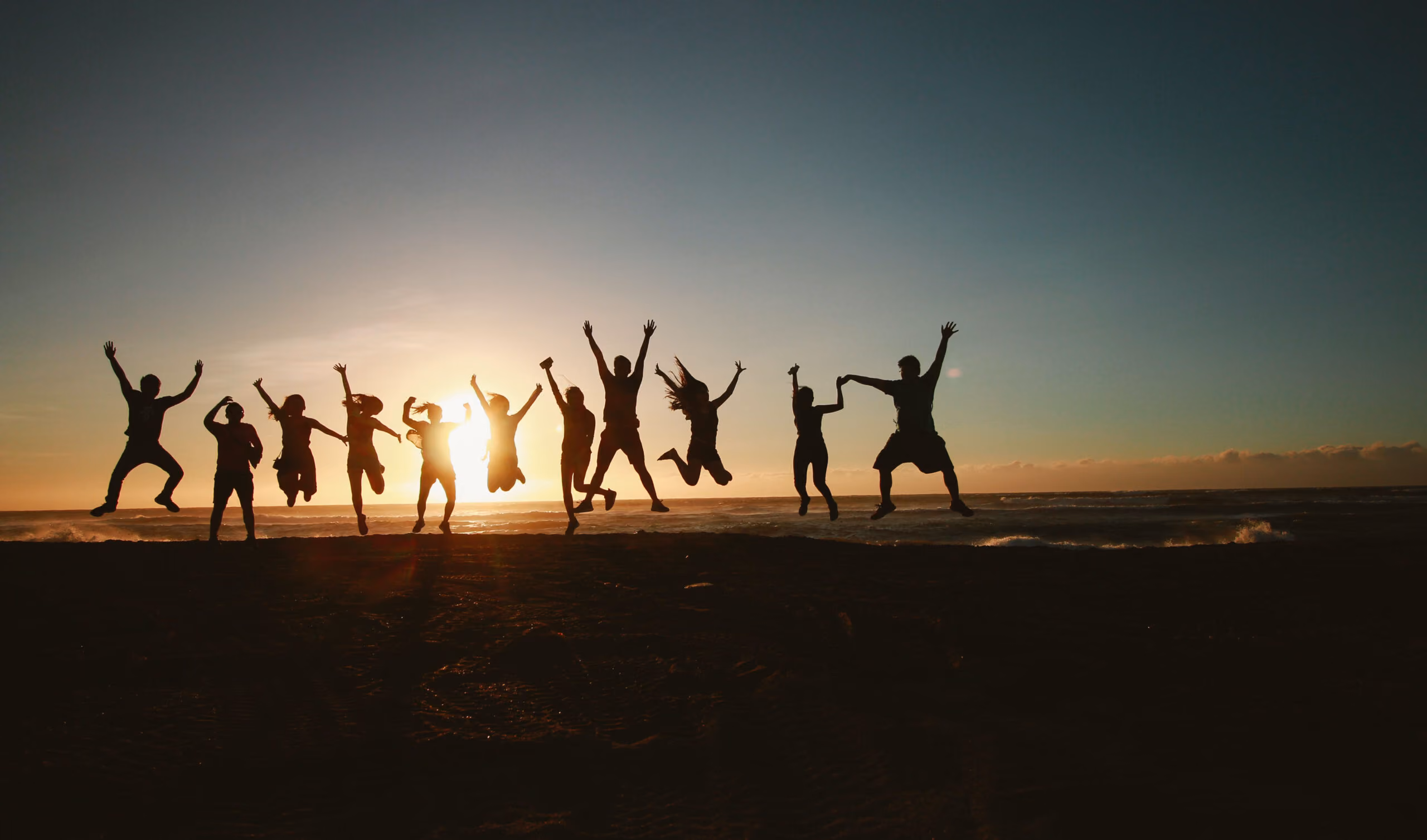 Gruppe von Menschen springt am Strand vor der untergehenden Sonne in die Luft.