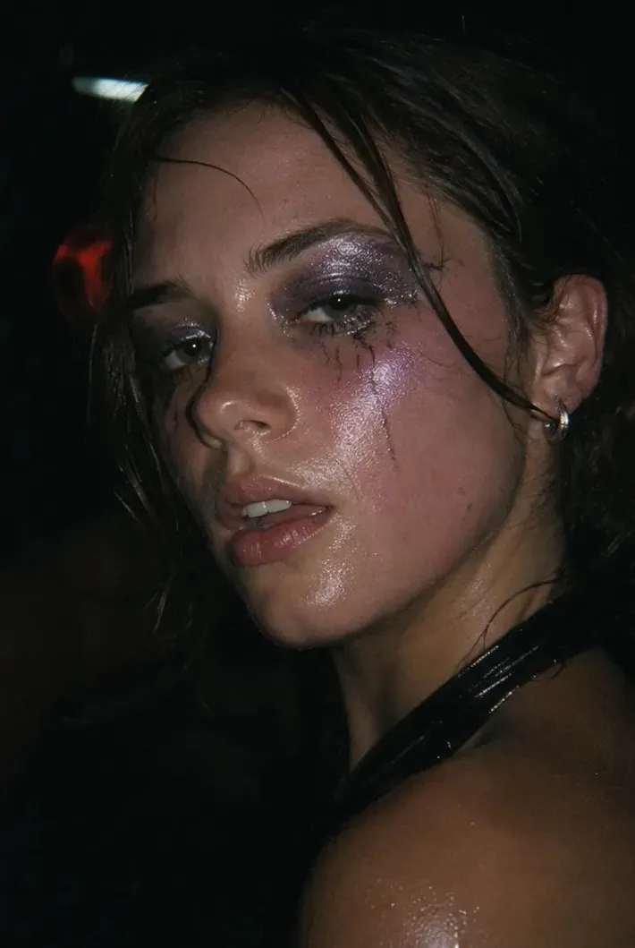 Close-up night portrait of a young woman with smudged purple glitter eye makeup and dewy skin under club light
