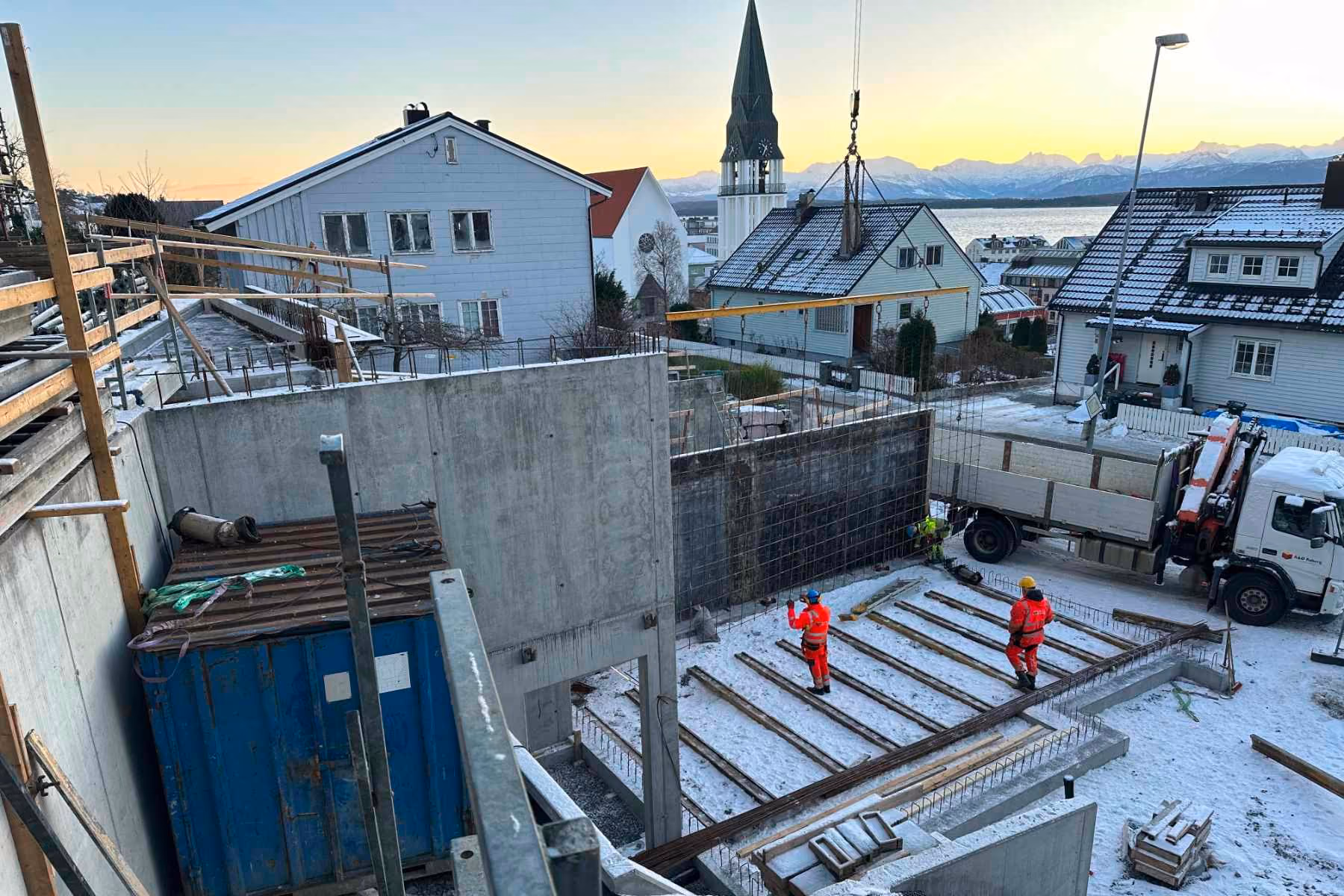 Construction site with workers in orange gear placing wooden beams on snow-covered ground near houses and mountains.