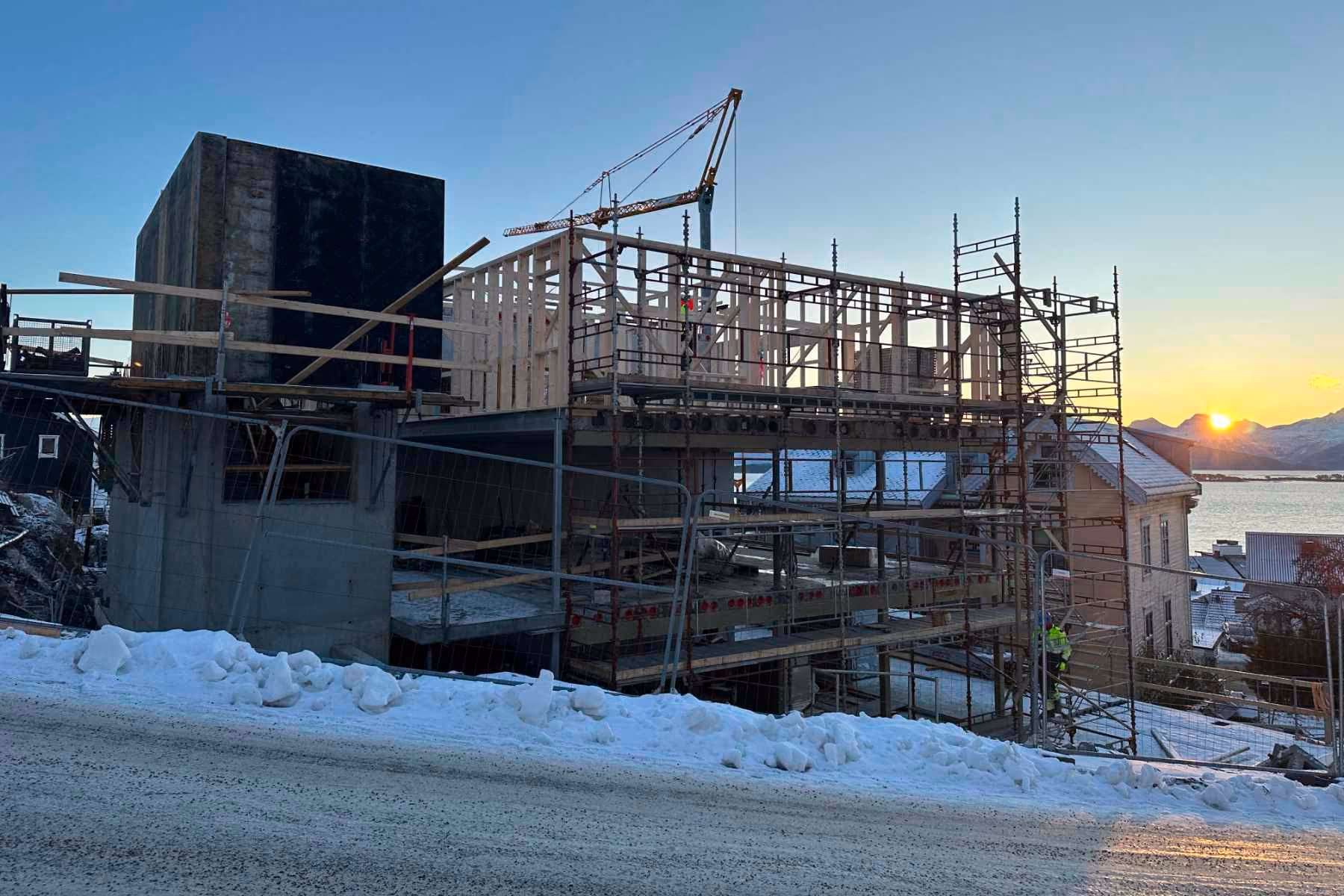Building under construction with scaffolding and wooden frame, snowy ground, and sunset over mountains.