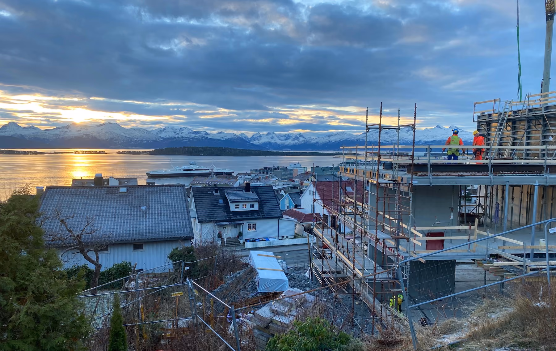 Sunset over snowy mountains, calm water, houses, and construction site with workers in safety gear.