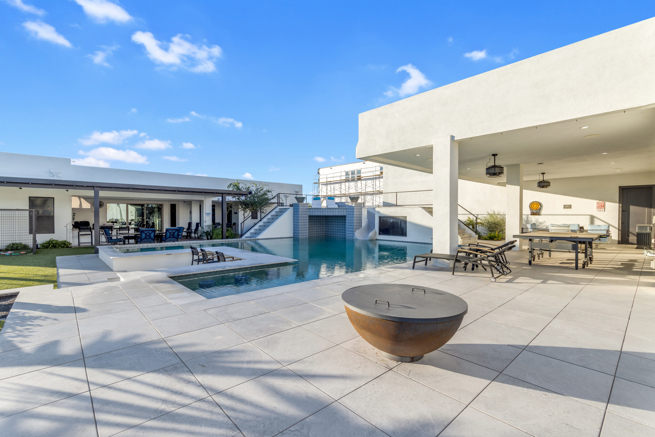 Modern outdoor pool area with lounge chairs, covered patio, fire pit, and clear blue sky.