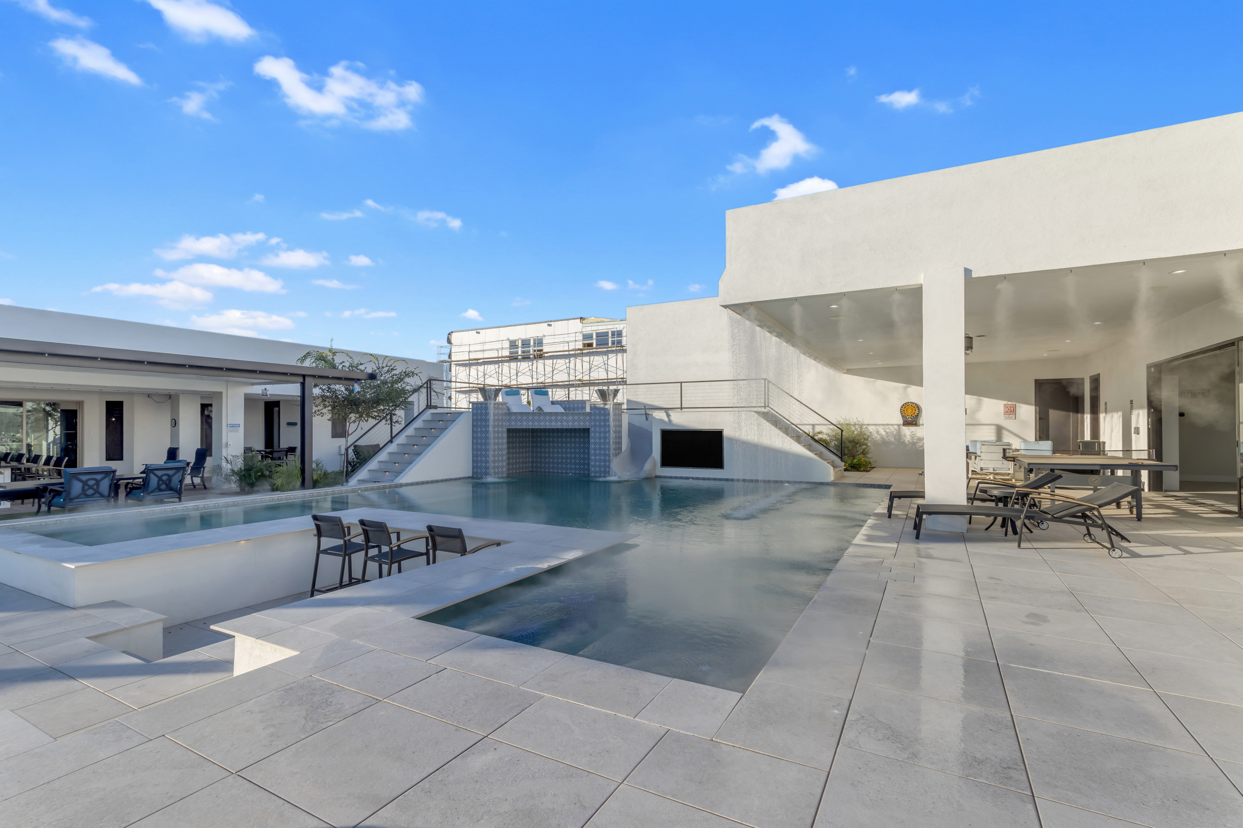 Modern outdoor pool area with sunken bar, lounge chairs, and covered patio with tables.