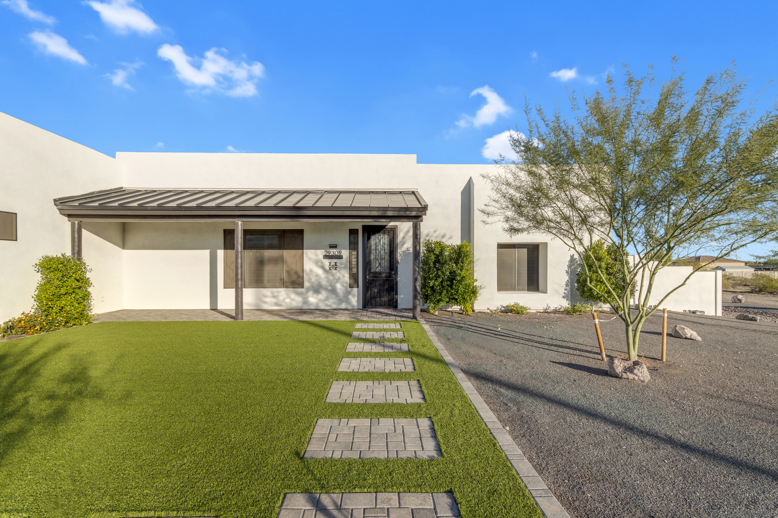 White modern house with a covered porch, green lawn with stepping stones, and a tree on a gravel yard.