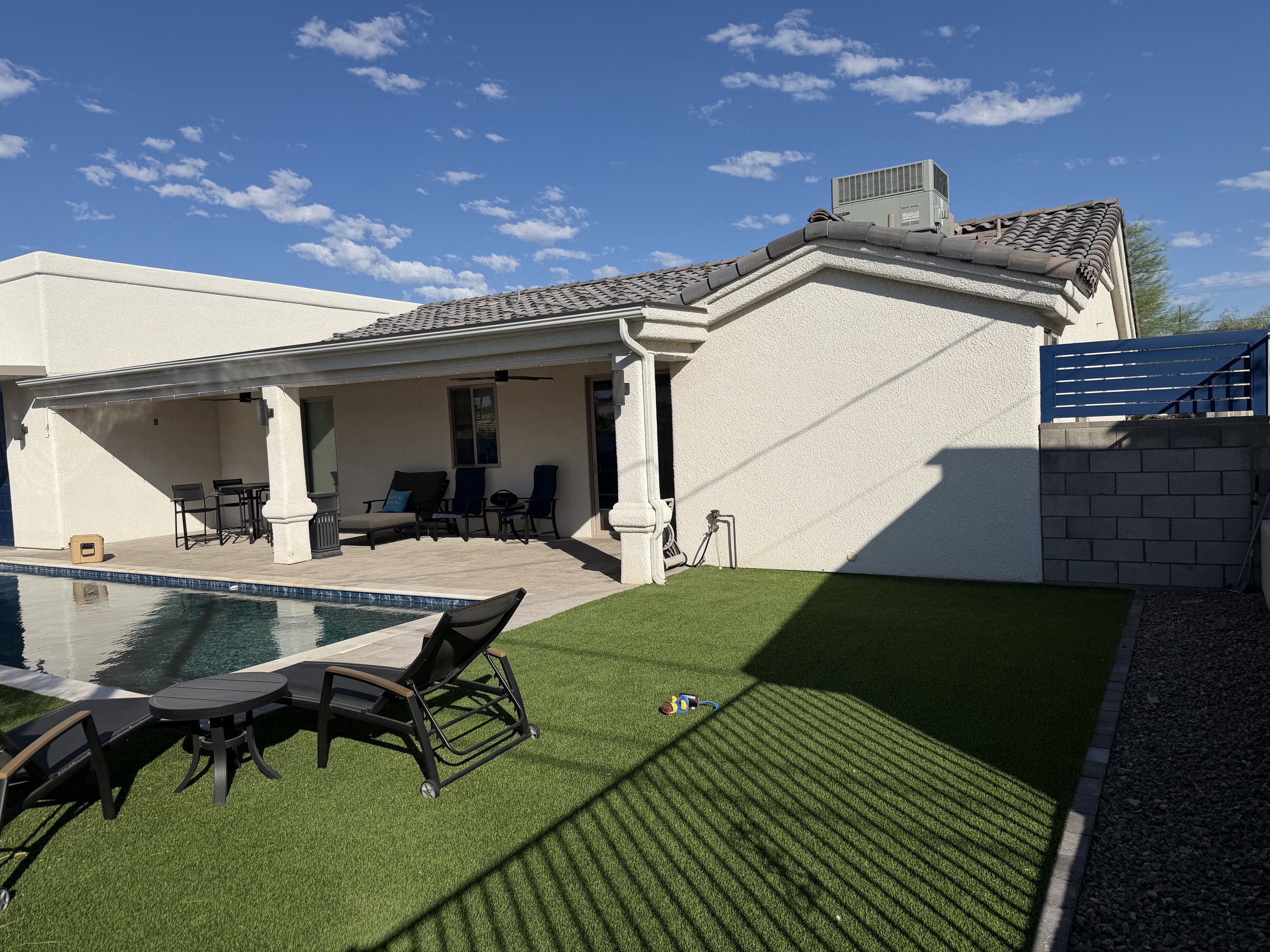 Backyard with pool, two black lounge chairs, small table, green artificial turf, and covered patio area.