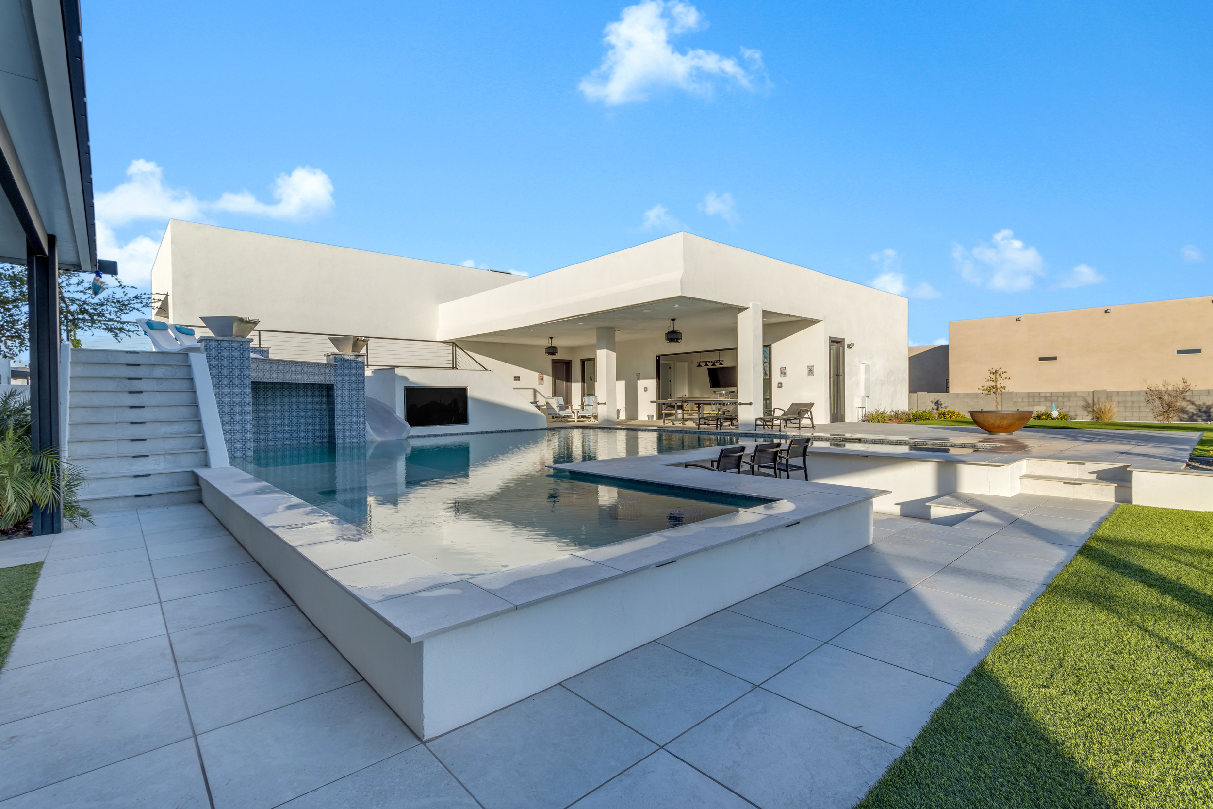 Modern backyard with infinity pool, outdoor seating, fireplace, and covered patio under blue sky.