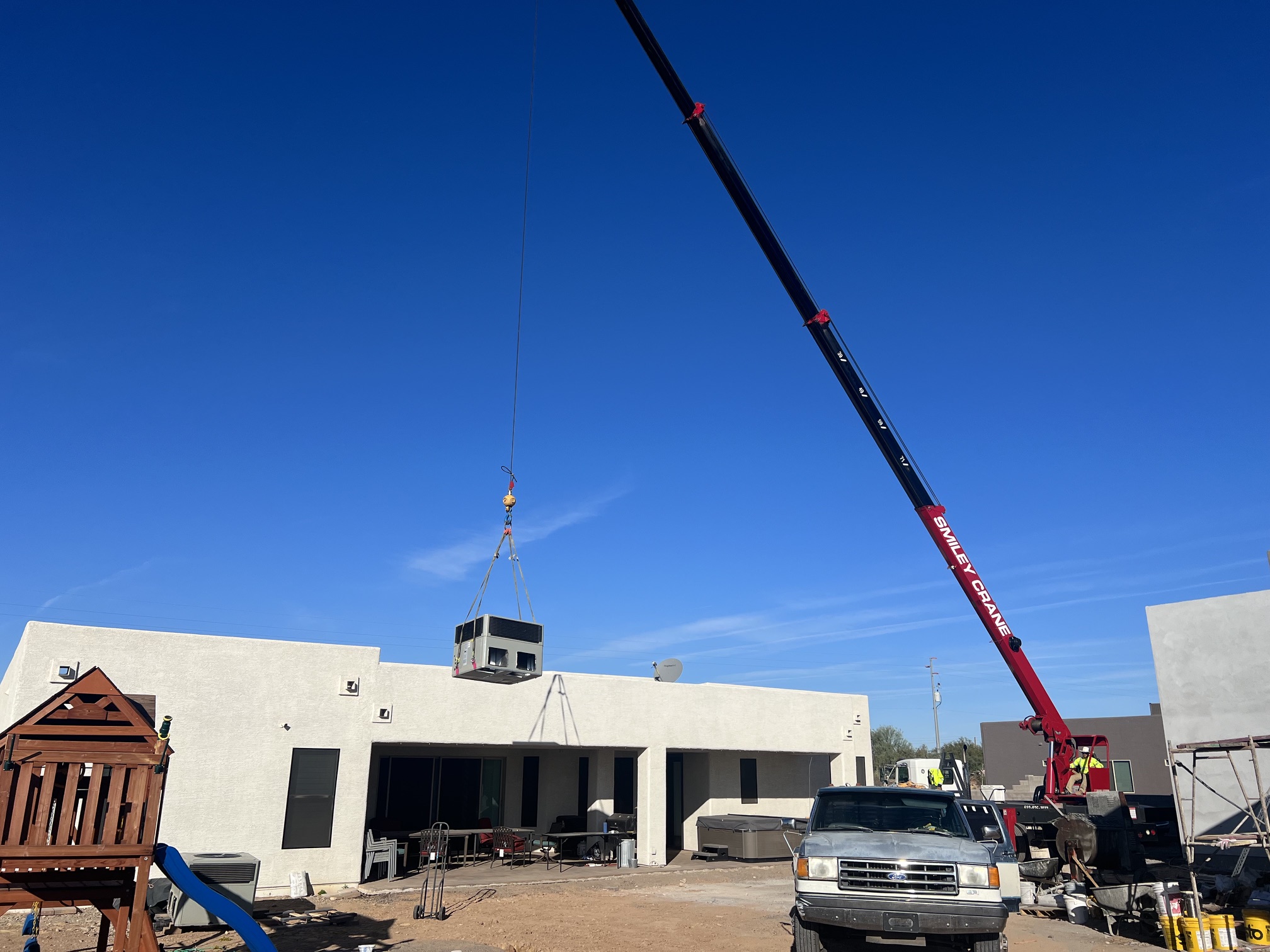 Crane lifting HVAC unit onto a flat-roofed building under clear blue sky with parked truck nearby.