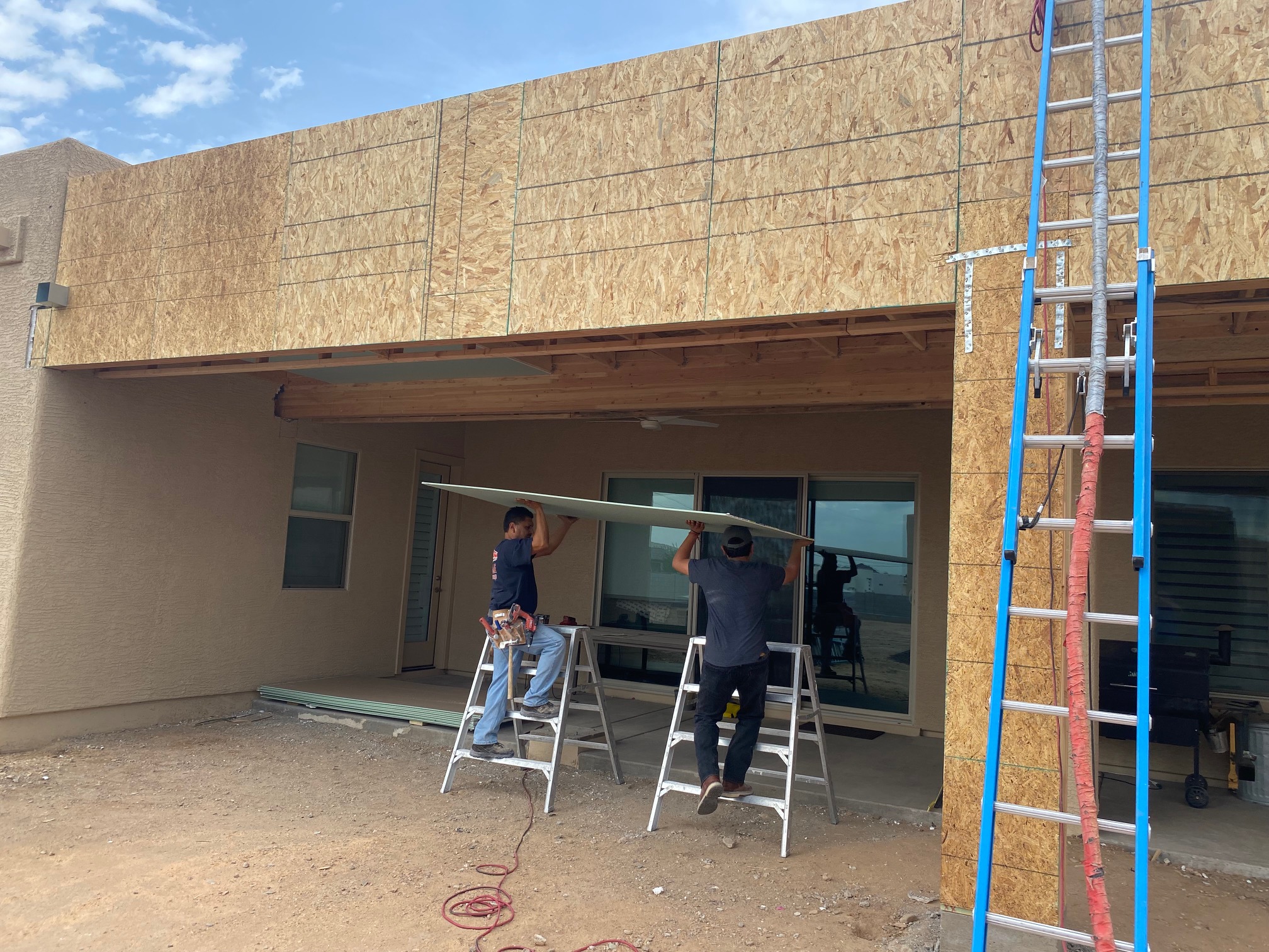 Two men on ladders lifting a drywall sheet in front of a building under construction.