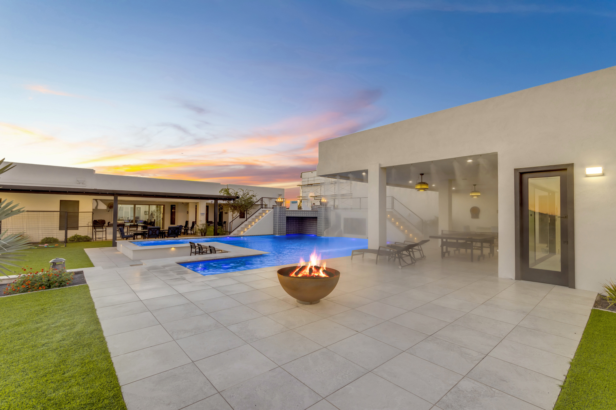 Modern backyard patio with a fire bowl, pool, lounge chairs, and covered seating at sunset.