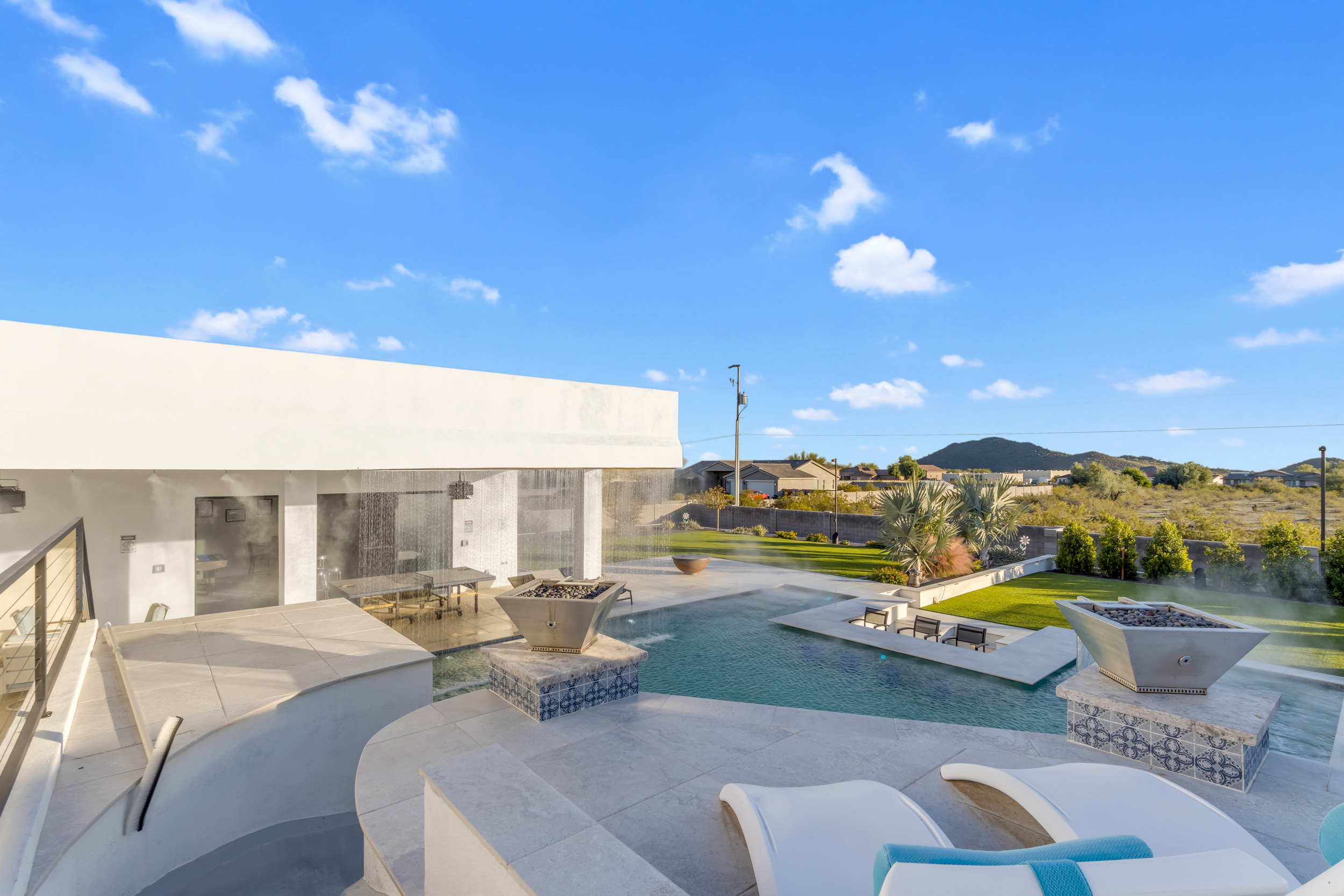 Modern home with swimming pool, waterfall feature, fire pits, and outdoor seating under clear blue sky.