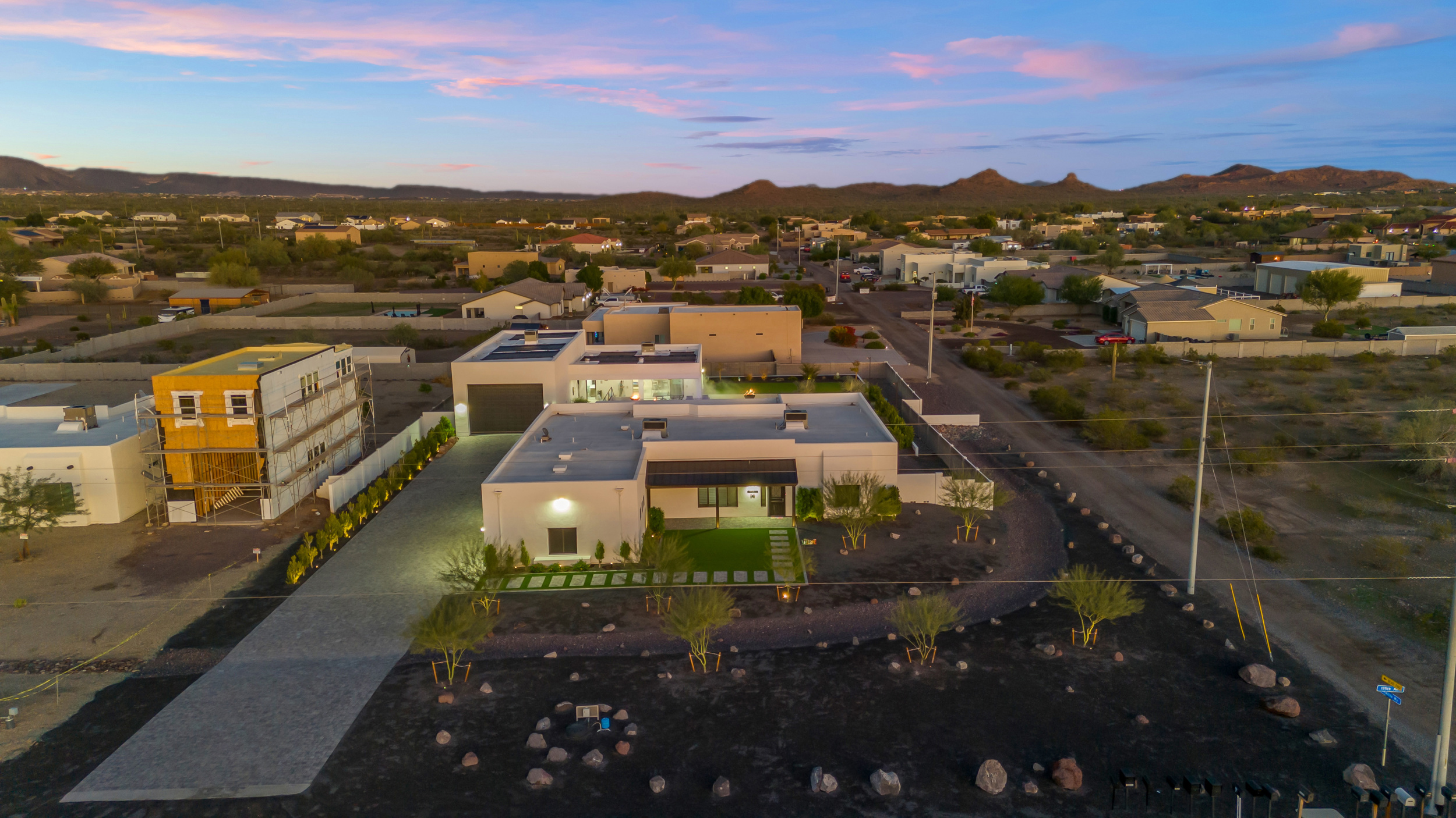 Aerial view of a modern white house with a long driveway in a desert neighborhood at sunset.