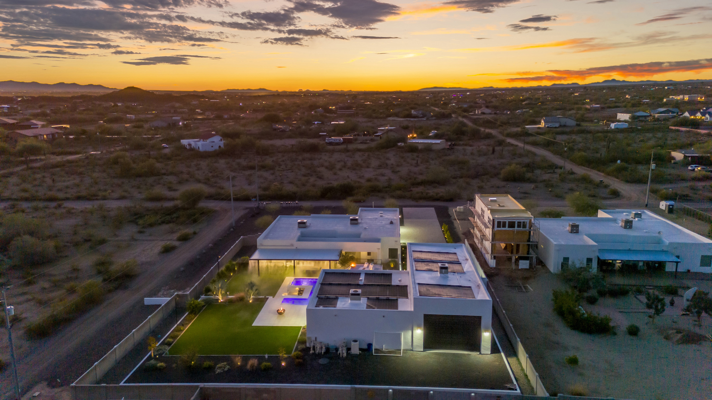 Modern white house with solar panels and pool lit up at dusk in a desert neighborhood.
