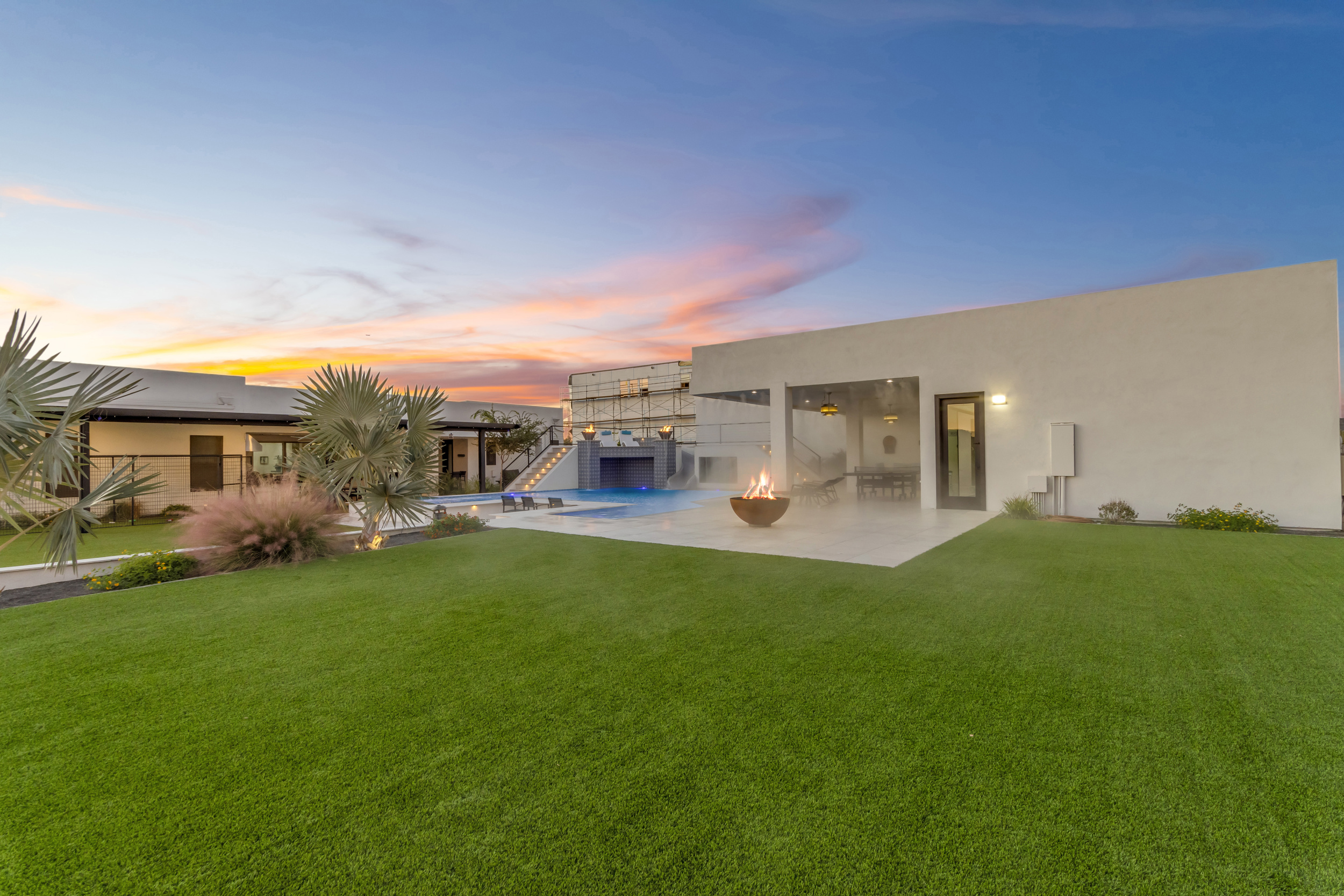 Modern backyard with green lawn, pool, fire pit, and patio at sunset under a colorful sky.