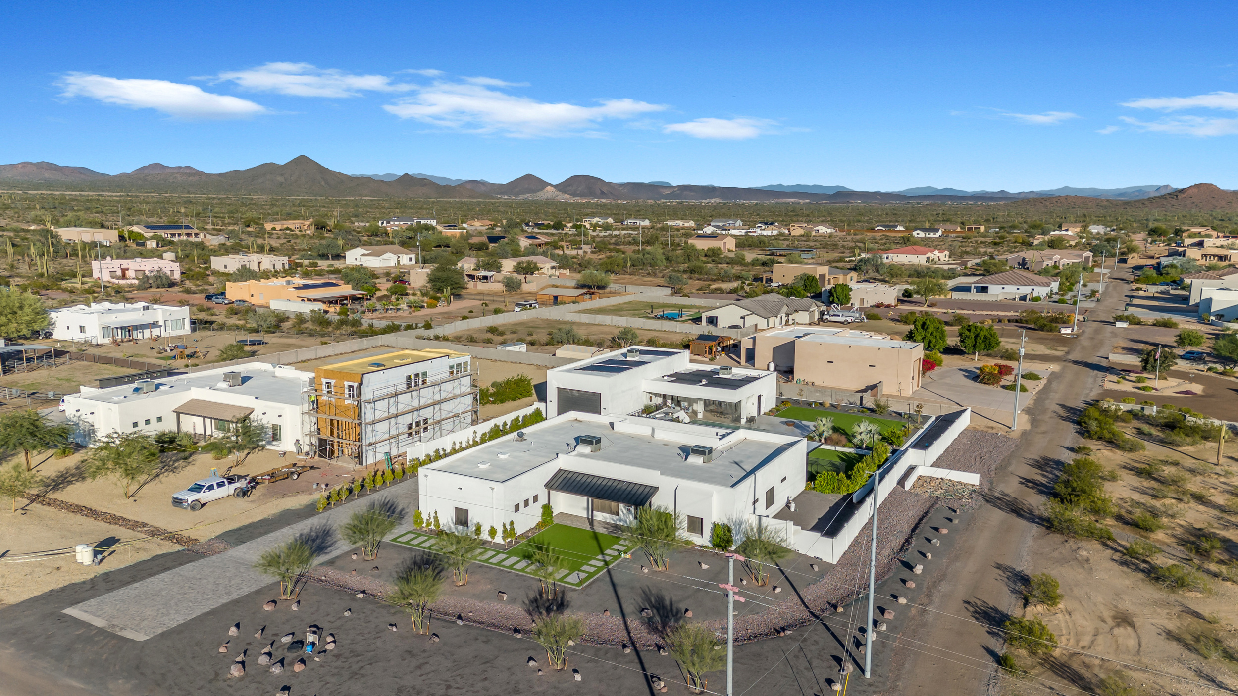 Aerial view of modern white desert homes with landscaped yards and mountainous horizon under clear blue sky.