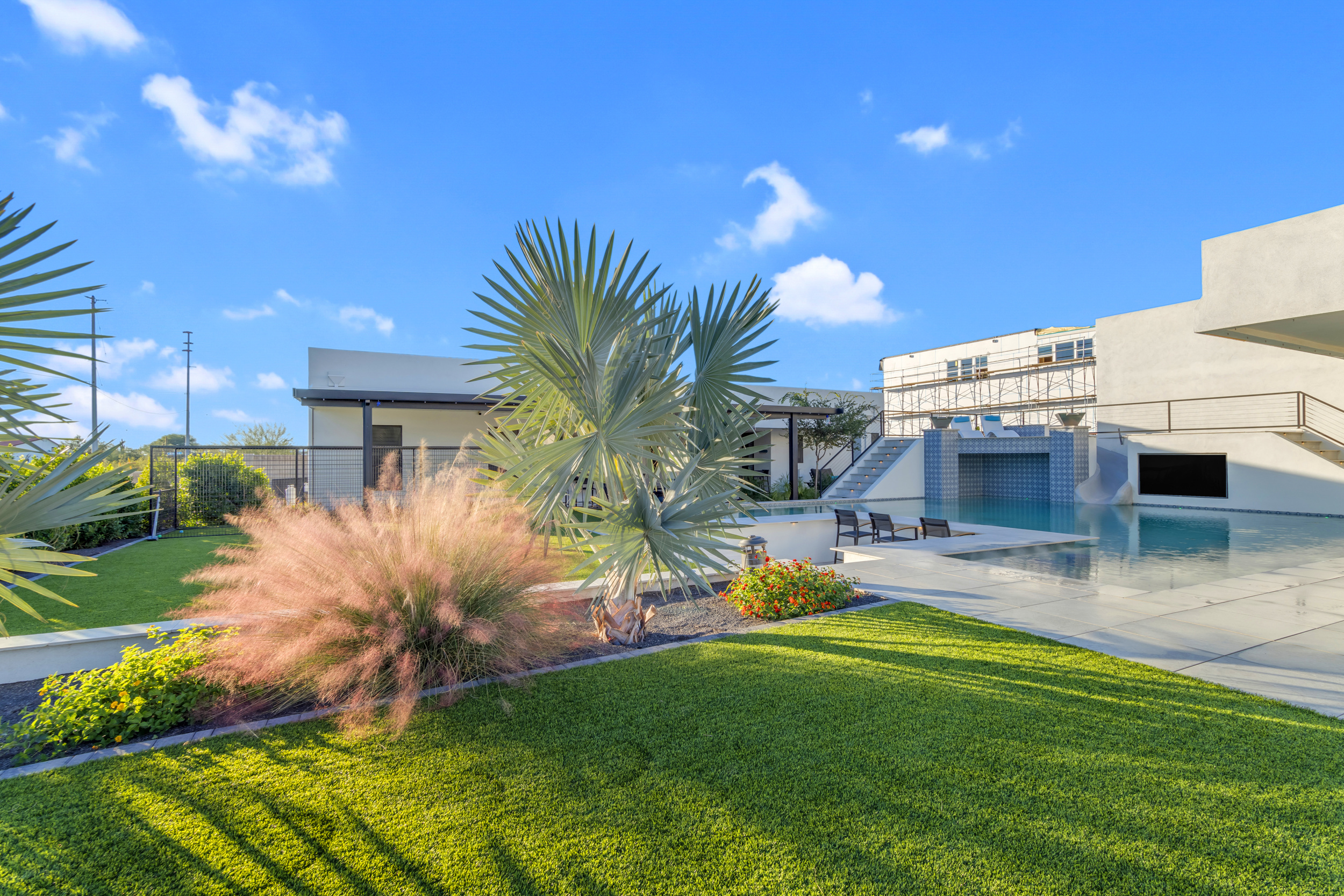 Modern backyard with green lawn, palm plants, patio area, and tiled pool under a blue sky.